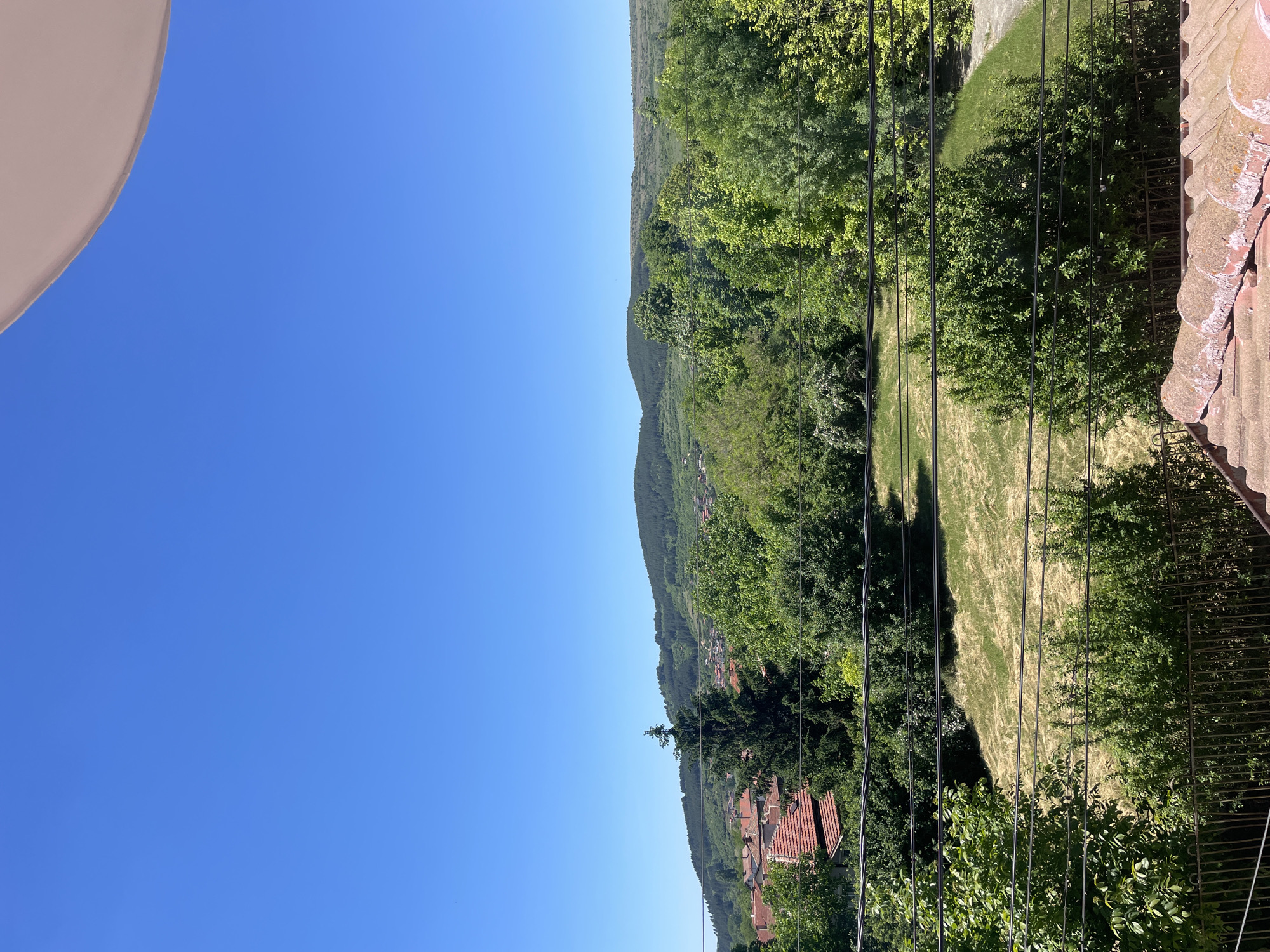 The image depicts a landscape with a clear blue sky occupying the left half of the frame. A mountain range covered in green vegetation extends horizontally across the image, transitioning from a lower elevation with visible red-roofed buildings to a densely forested upper slope. Black overhead wires run vertically through the forest. In the foreground, on the right side of the image, there is a glimpse of a roof with red tiles and some foliage from nearby trees. The location is Yahinovo, Bulgaria.