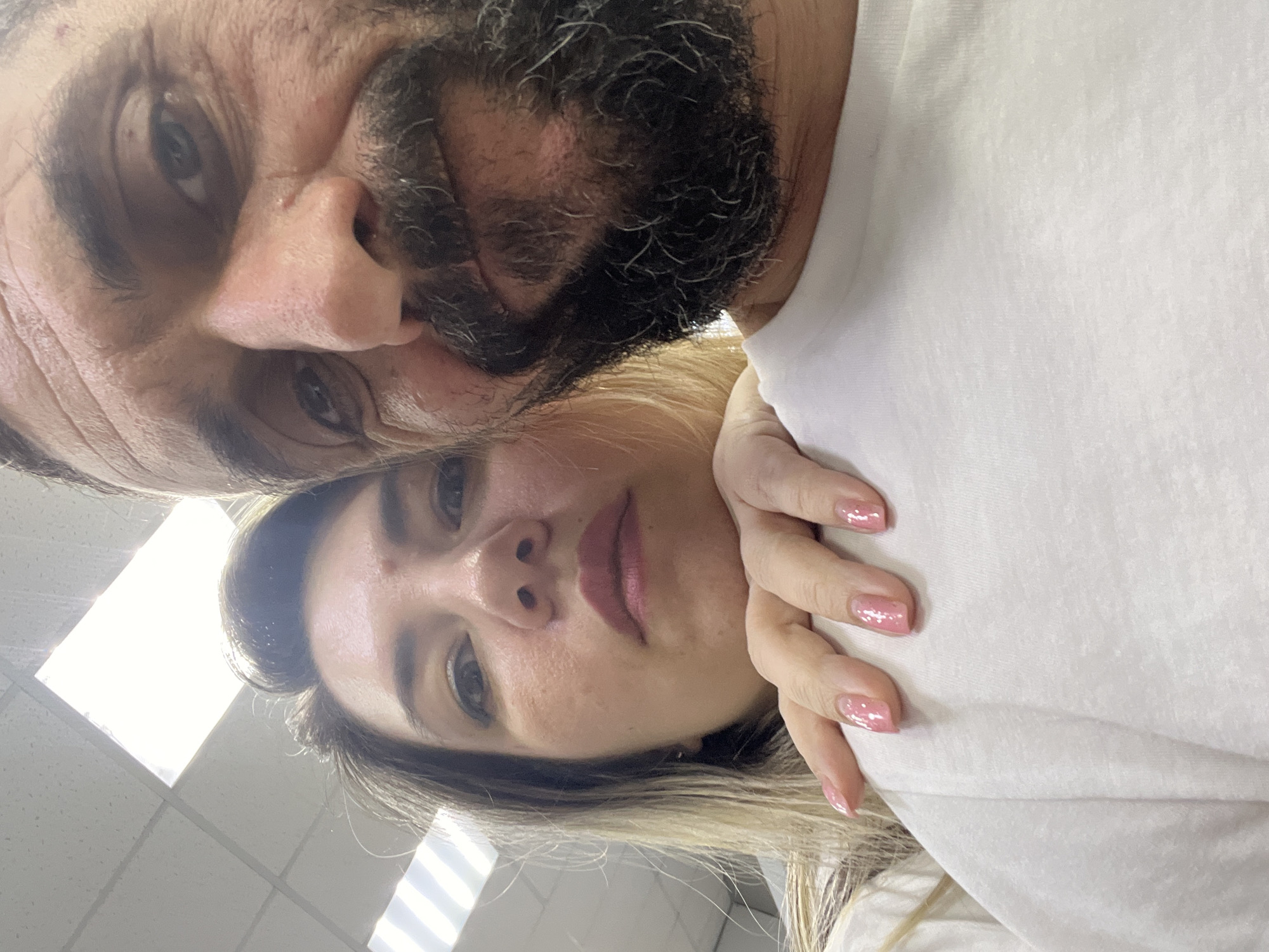 A close-up, low-angle selfie shows a man and a woman. The man, on the right, is in the foreground, with his face taking up a significant portion of the frame. He has a dark, grizzled beard and is looking directly at the camera with his eyes open. The woman is positioned behind and to the left of the man, her face visible from the nose up. Her hand rests on the man's shoulder, and her nails are painted a shimmery pink. Both individuals are wearing white shirts. The background appears to be an indoor space with a white tiled ceiling that has fluorescent lighting panels. The location is Unknown, Unknown.