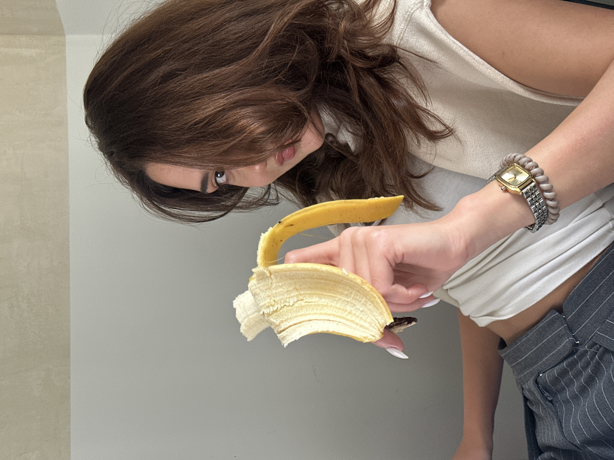 A woman with brown hair is inspecting a peeled banana in her right hand. A snake, partially hidden inside the banana peel, is visible. The woman is wearing a white top, grey striped pants, and a gold wristwatch with a beaded bracelet. The background is a plain grey wall. The image doesn't provide any information about the location being in Yerevan, Armenia.