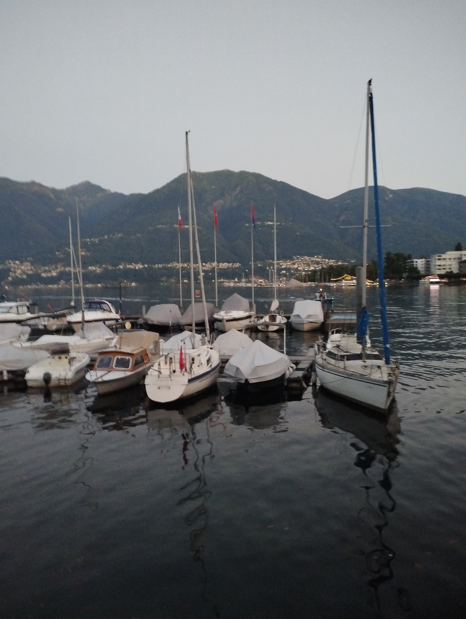 The image depicts a marina in Muralto, Switzerland, with several boats moored on a calm body of water. The sky is overcast, and the surrounding landscape features rolling hills and mountains. In the foreground, a white sailboat with a blue sail is docked. Other boats of various sizes, some covered with tarps, are also visible. The water reflects the boats and the overcast sky. The scene appears to be captured during twilight or early evening.