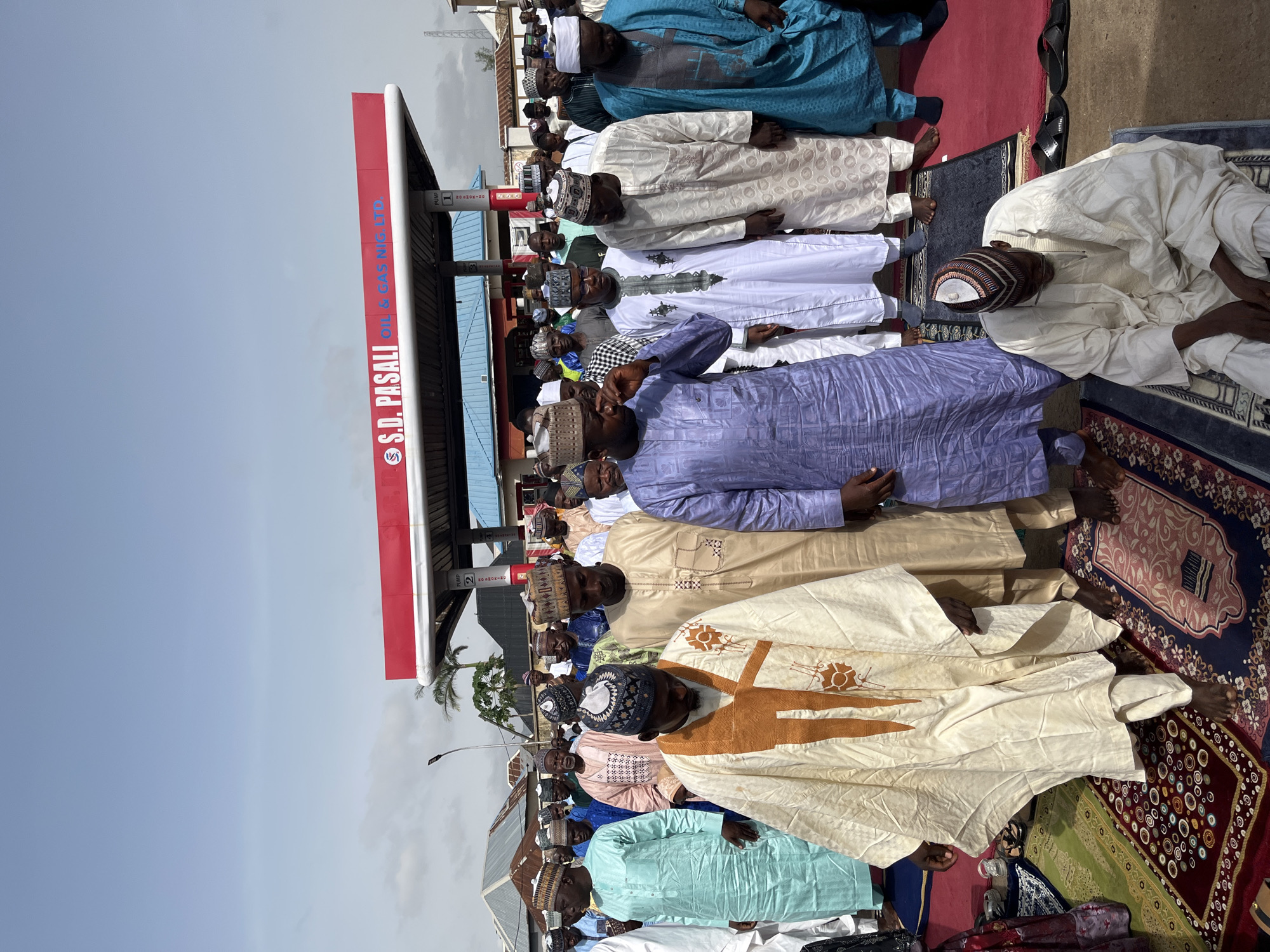 A group of men are praying outside at an S.D. Pasali Oil & Gas Nig Ltd. gas station in Jos, Nigeria. They are wearing various colored robes and hats. Some men are standing while others are kneeling on prayer rugs.