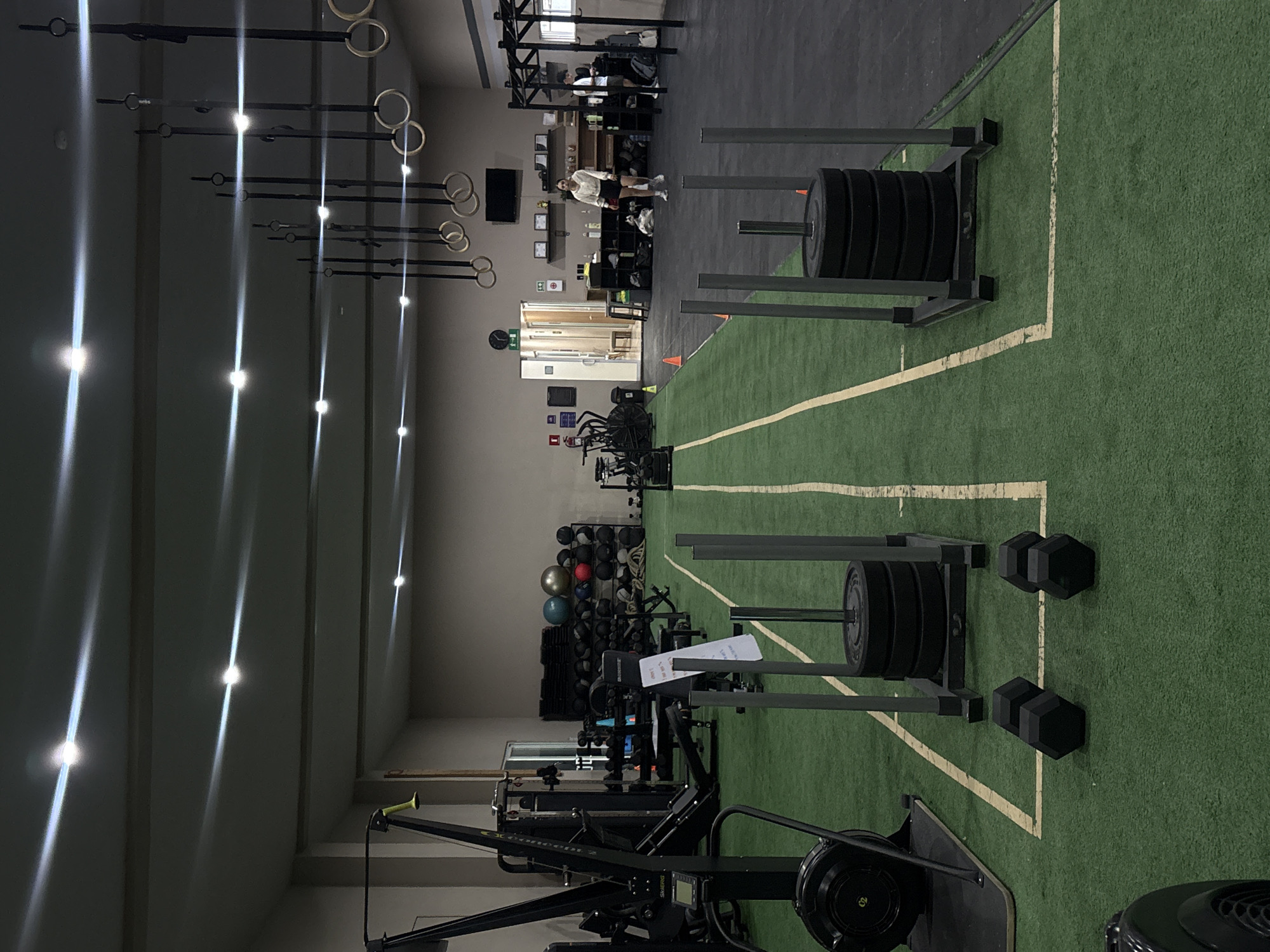 The image depicts the interior of a fitness center. The location is stated to be in San Bernardino Tlaxcalancingo, Mexico, although this cannot be verified from the image itself. The gym features various exercise equipment including weight sleds loaded with plates, dumbbells, rowing machines, and a rack of medicine balls. There is green artificial turf with white lane markings on the floor. Suspended from the ceiling are gymnastic rings. A woman stands in the background near a weight rack, wearing a light-colored shirt and shorts. The lighting comes from overhead recessed lights. The walls are a neutral color.