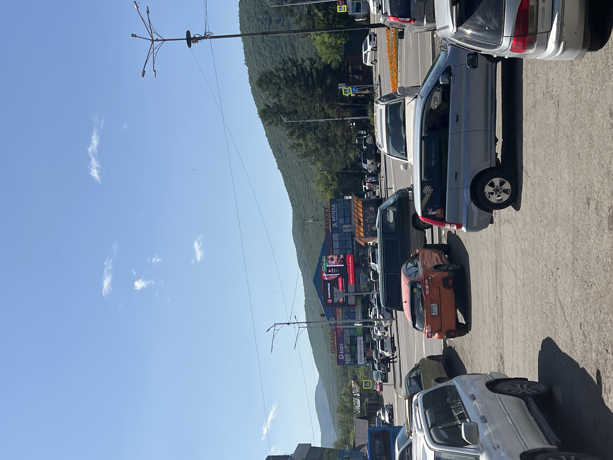 In Yelizovo, Russia, on a clear day with a bright blue sky and scattered clouds, a multi-lane street is filled with traffic. A white SUV is parked in the foreground on the left. Further down the street, an orange hatchback is stopped at an intersection. To the right of the orange car, a light blue minivan is also in traffic. In the background, several commercial buildings display signage in Cyrillic, including "Kari" and "Apteka," indicating retail establishments. Beyond the buildings, a lush green mountain range rises, covered in trees. Streetlights and power lines are visible above the road. Several pedestrians are crossing the street at a designated crosswalk, accompanied by a traffic sign. The scene captures a typical urban environment with vehicular activity and commercial infrastructure against a natural mountainous backdrop.