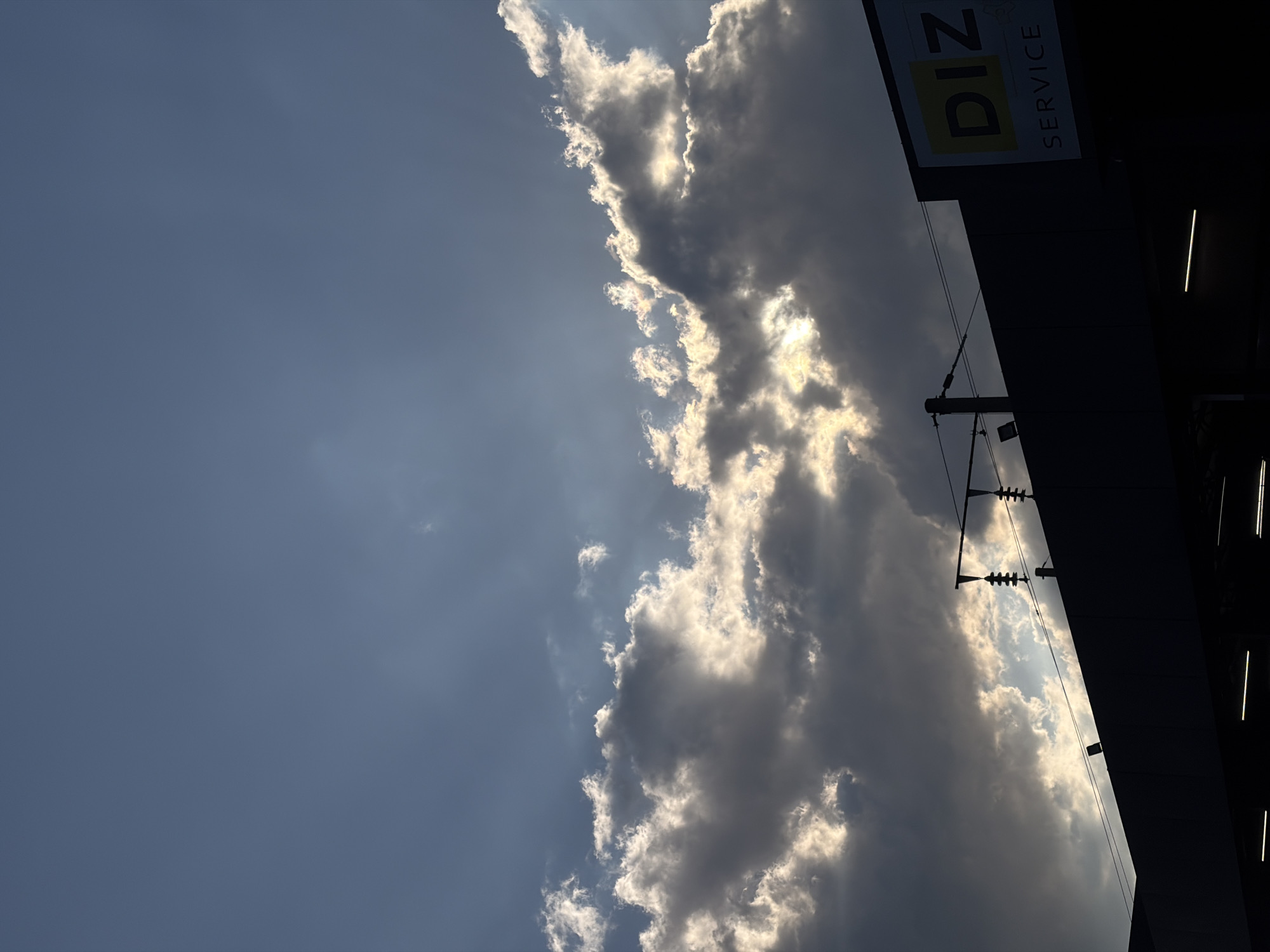The image, taken outdoors, features a partially cloudy sky beside a building. The left side of the image shows a blue sky with some thin white clouds. The right side is dominated by dense, dark grey clouds, with sunlight filtering through some gaps. The edge of a building can be seen on the right side. The sign "DIZ SERVICE" is visible on the building. Given the information, the location is likely in Krasnodar, Russia. The scene is generally still, with the clouds being the main dynamic element.