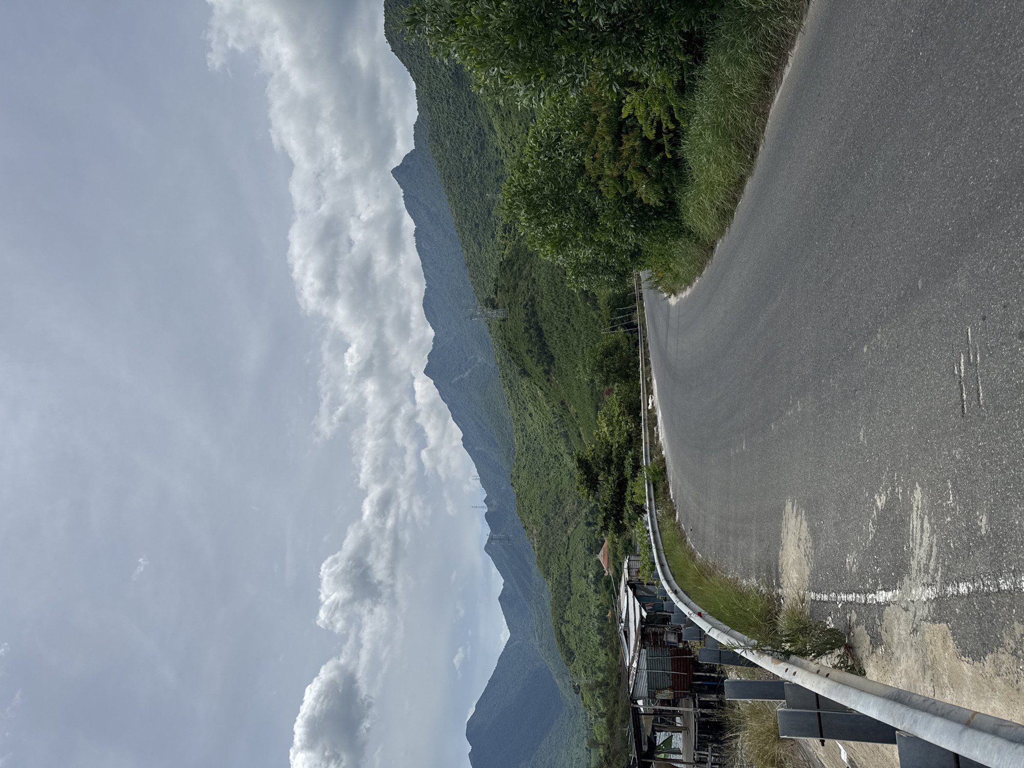 A paved road curves through a lush, green mountainous landscape under a cloudy sky. On the left side of the road, there are some dilapidated structures built on stilts, appearing to be houses or shacks. A metal guardrail runs along the edge of the road. The mountains in the background are covered in dense vegetation. Power lines and transmission towers are visible on the slopes of the mountains. The scene suggests a rural or semi-rural area near the outskirts of Huế, Vietnam, possibly along a coastal or riverside road. The overall atmosphere is one of natural beauty combined with signs of human habitation.