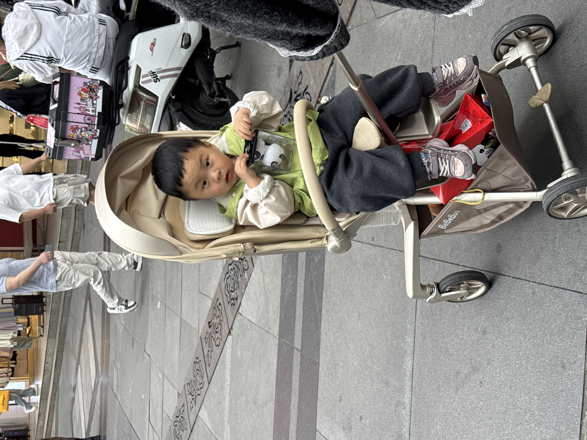 A toddler in a stroller, wearing a green jacket and gray pants, holds a toy, positioned on a gray tiled sidewalk with a patterned inset; Jinjiang District, China. Pedestrians walk on the sidewalk behind the stroller, and a white motorcycle is parked nearby. Red boxes are stowed in the stroller's storage area.