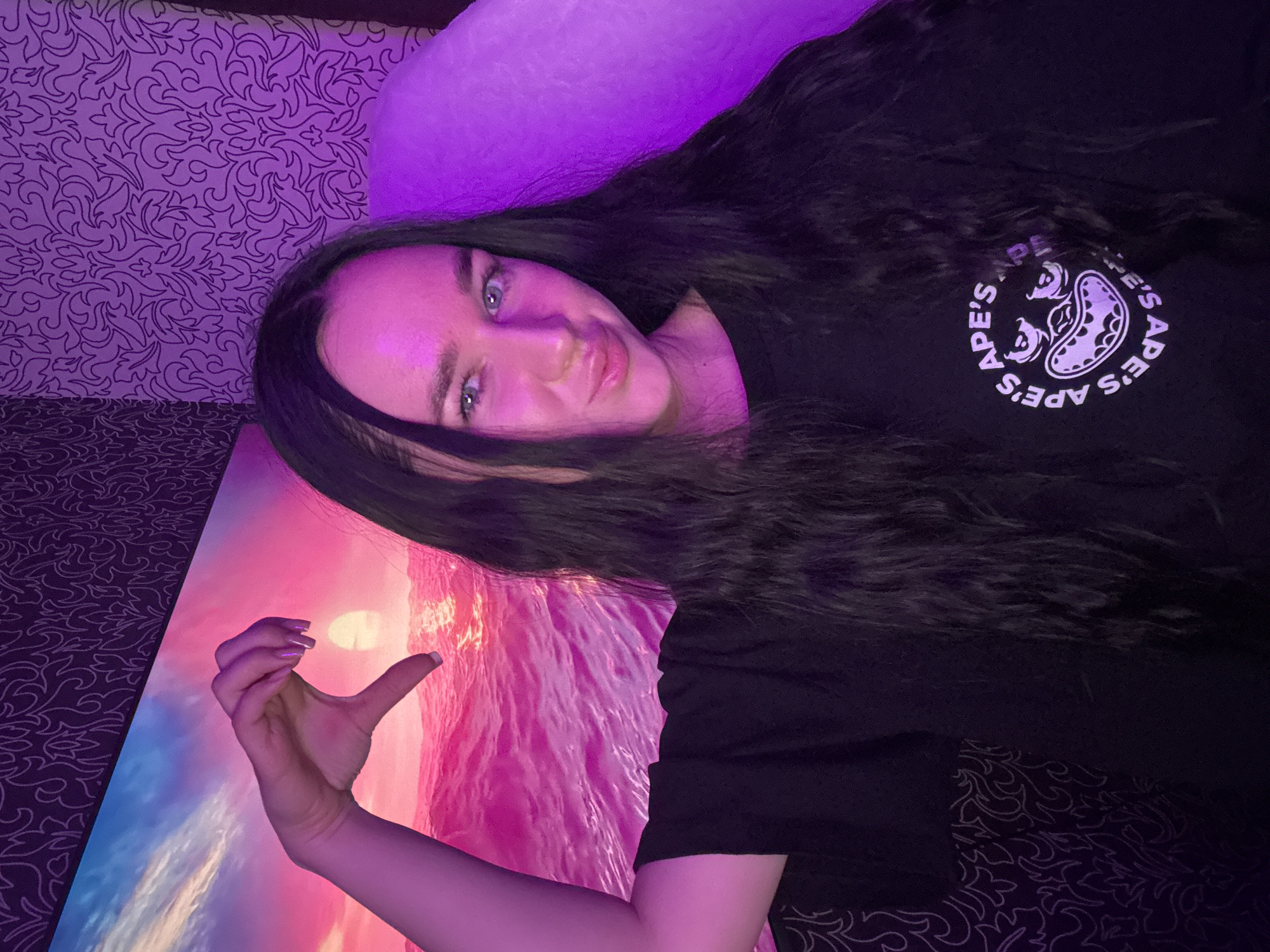 The media file depicts a young woman lying down, with her head propped up on a pillow. She has long, dark hair and is wearing a black t-shirt with a circular logo featuring ape faces. The scene is illuminated by purple lighting, casting a vibrant glow over the woman and the surrounding area. To her left, a large, colorful artwork is visible, showcasing a sunset over a body of water, with dominant pink and orange hues. Her right hand is held up in front of the artwork, forming a heart shape with her thumb and index finger, with her fingernails painted in a light purple shade. The background appears to be a patterned textile, possibly a bedspread or wallpaper, with a dark base and a lighter, intricate design. The overall mood is relaxed and intimate, likely captured indoors given the lighting and the artwork. There is no visible text on the artwork or the woman's clothing apart from the logo on her shirt, which reads "APE'S APE'S".