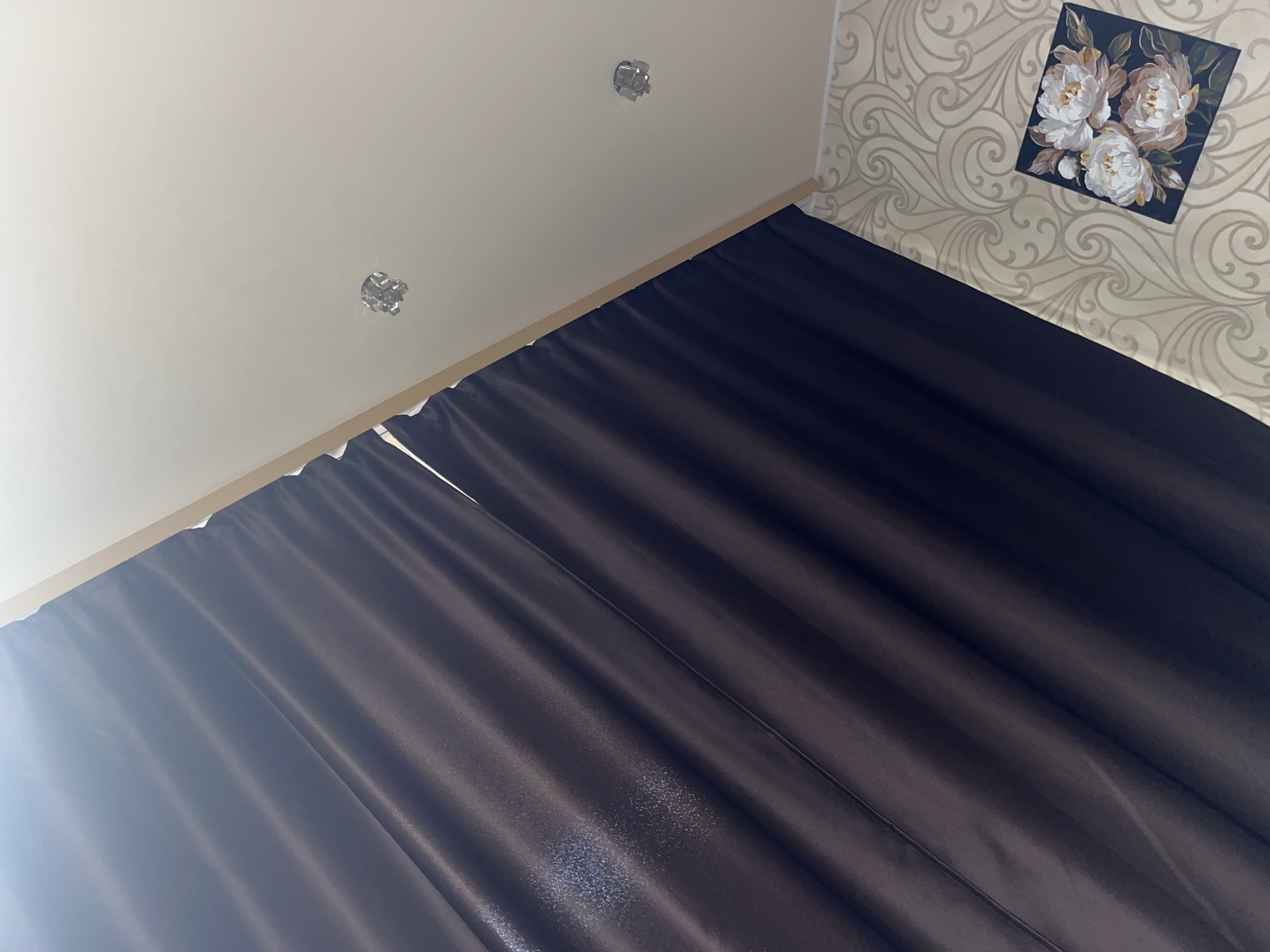 The image displays an interior scene with a dark brown curtain dominating the left side. The curtain is pleated and appears to be made of a satin-like material, reflecting light in some areas. The right side of the image shows a white sloped ceiling with two circular light fixtures. Below the ceiling, there is a section of wallpaper with an intricate swirling pattern in a light beige color. In the bottom right corner, a small square artwork depicting three light-colored peonies on a dark background is visible. The scene appears to be a residential interior, potentially a living room or bedroom. There is no indication of a specific location in Chelyabinsk, Russia.