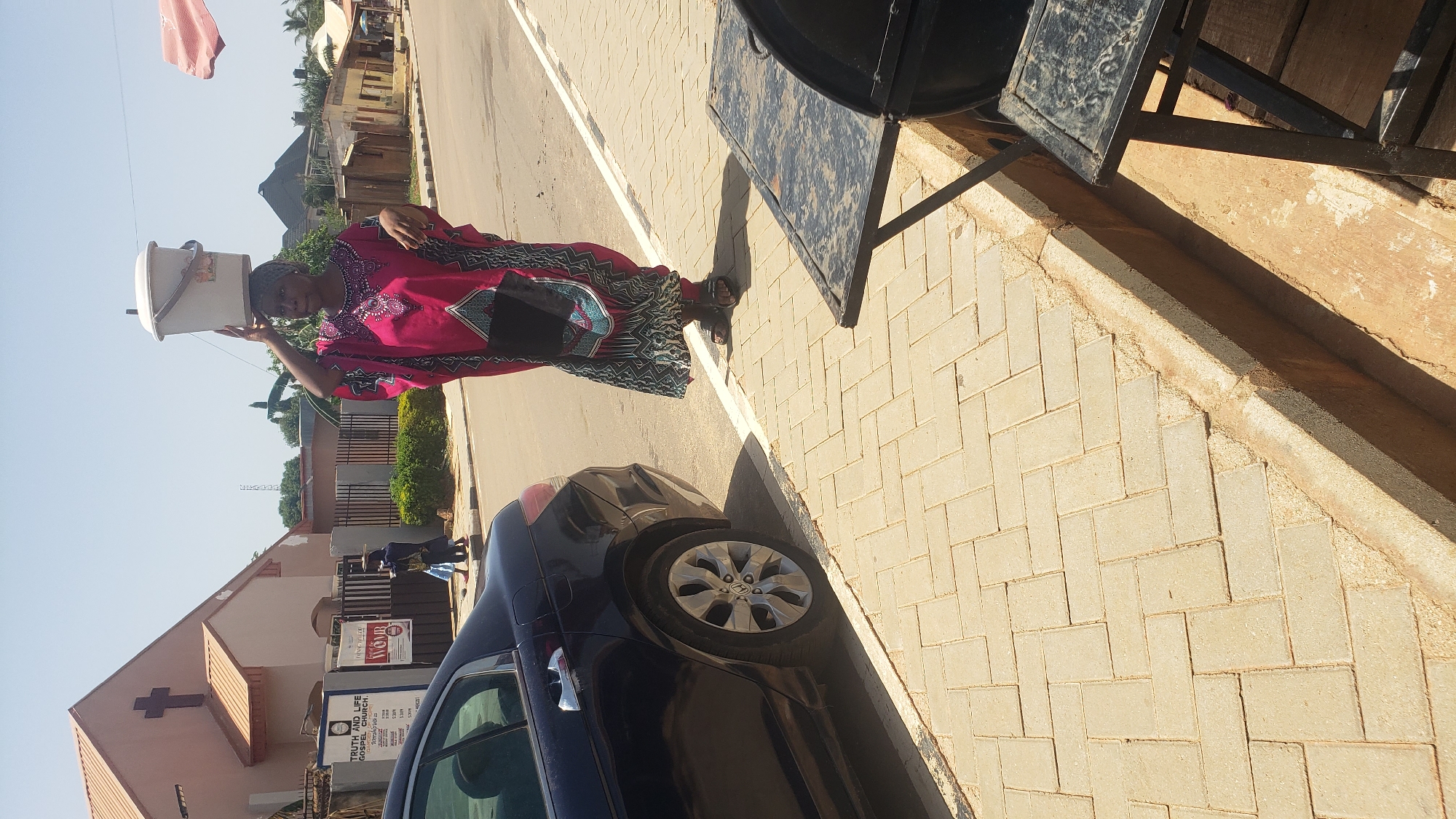 A woman balances a white bucket on her head as she walks down a paved street in Narayi, Nigeria. She wears a vibrant pink and black patterned kaftan and dark sandals. To her left, a dark blue car is parked on the side of the road. In the background, a light-colored church building with a cross on its roof is visible, along with other buildings and vegetation. The day appears to be sunny and clear.