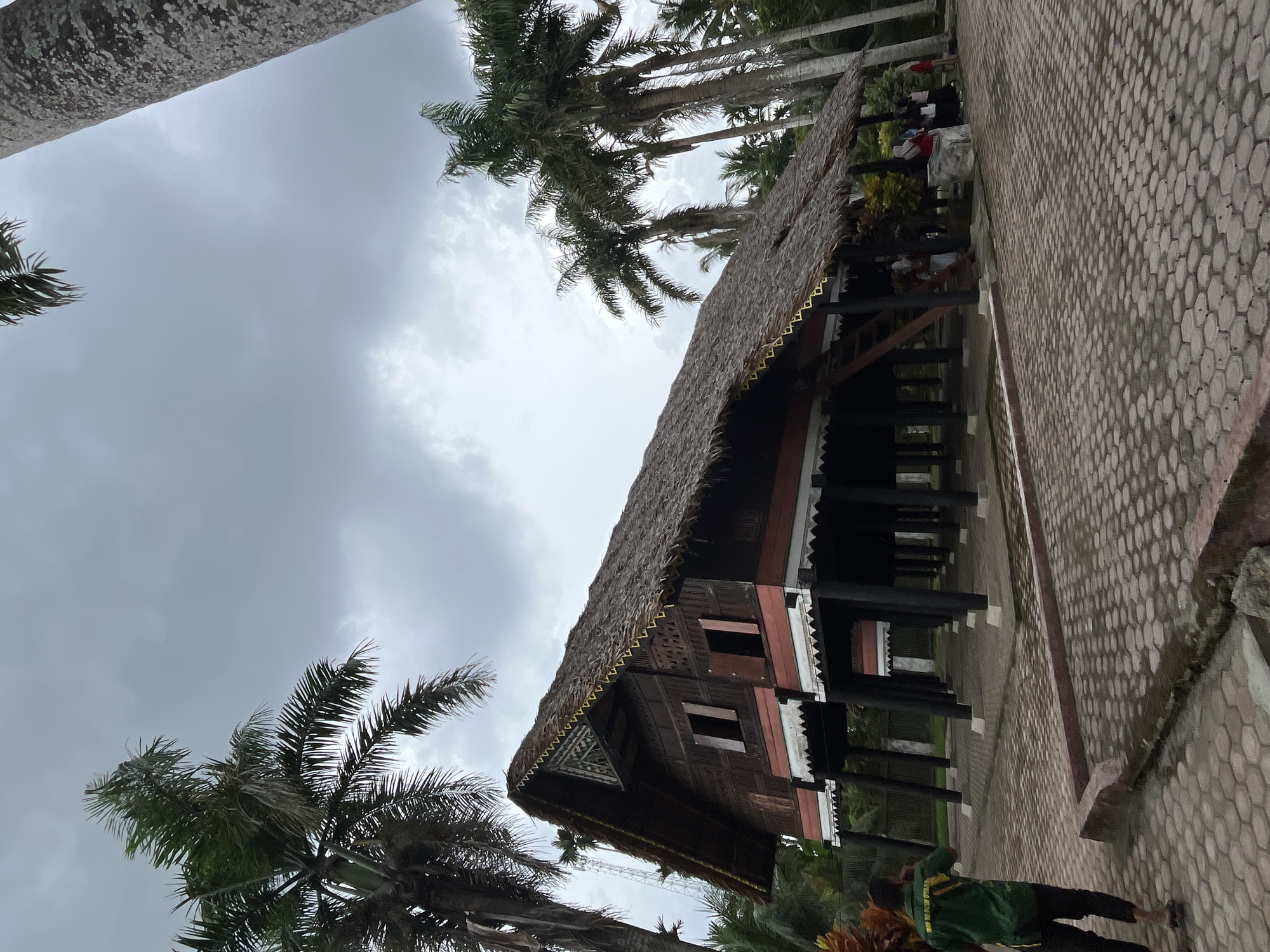 In the city of Mesjidpirak, Indonesia, an image captures a traditional stilt house with a thatched roof, surrounded by lush palm trees under a cloudy sky. The house, constructed of dark wood with red accents and white decorative trim, stands on numerous dark, slender pillars. A wooden staircase leads up to an open entrance on the side of the house. The ground around the house is paved with hexagonal interlocking stones, bordered by a low, reddish-pink concrete curb. A person in a green shirt and dark pants is seen walking away from the camera on the left. Several other individuals are standing near the house on the right, some appearing to be tourists or visitors. The overall scene depicts a cultural heritage site within a tropical environment.