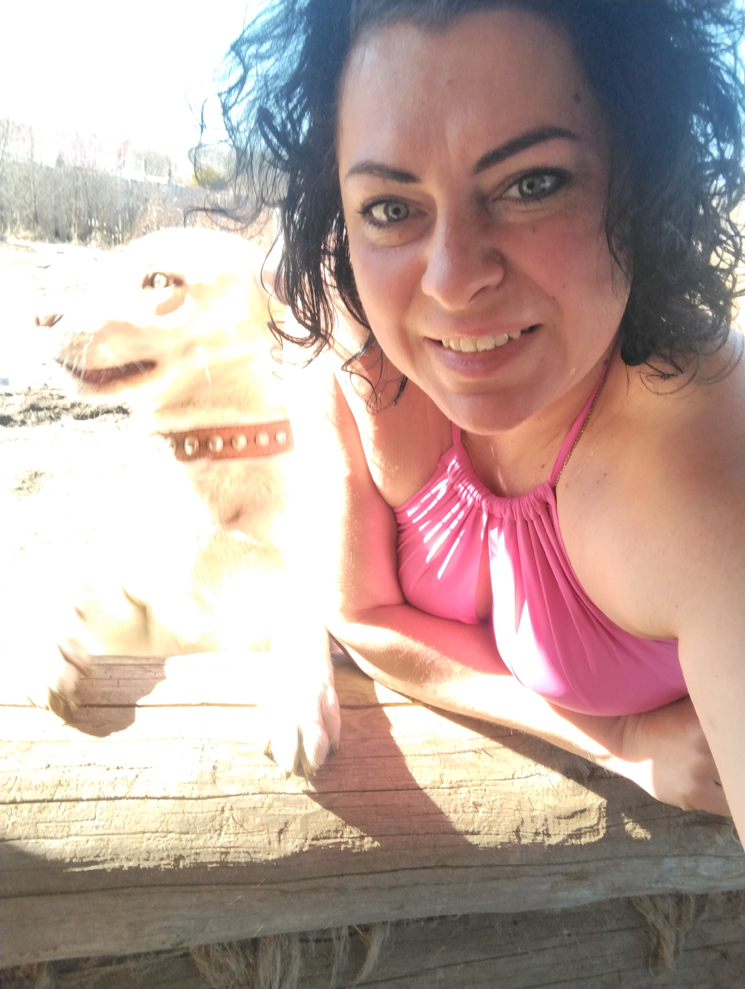 A selfie of a woman with curly dark hair, fair skin, and wearing a pink swimsuit. She is smiling and lying on a wooden surface. Beside her is a pale yellow dog wearing a studded collar. The background is blurred, appearing to show trees and vegetation. The location cannot be determined from the image. The image appears to be taken outdoors in bright sunlight. There is no reliable indication this image was taken in Grishanki, Russia.