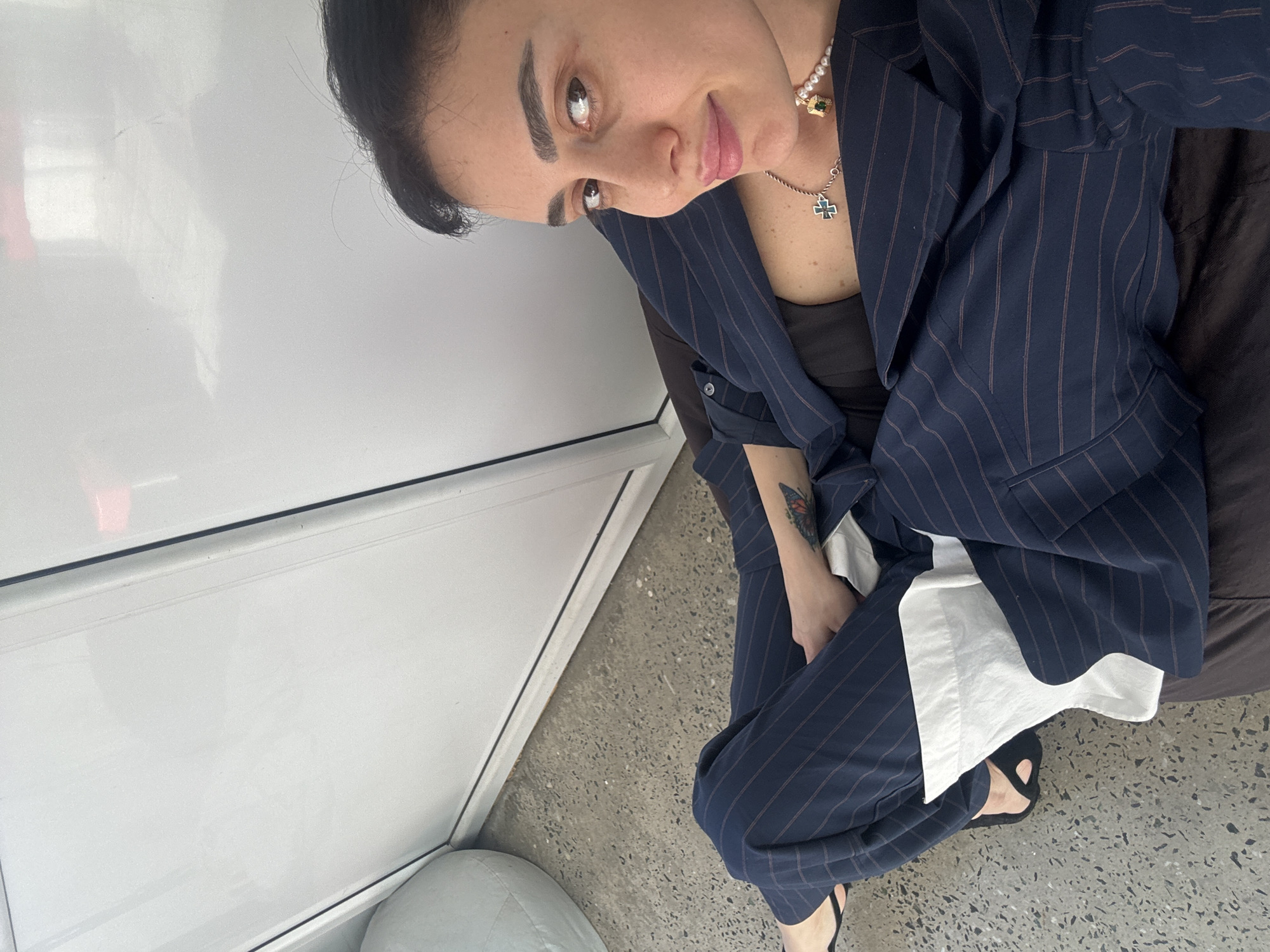 An eye-level shot features a woman seated indoors, looking directly at the camera with a subtle smile, likely in Tbilisi, Georgia. She has dark hair and pronounced eyebrows. She is wearing a navy blue pinstripe suit, comprising a jacket and trousers, over a black top and a white undershirt.
Her accessories include a pearl necklace with a green gem charm and a silver-toned necklace with a cross pendant. A colorful butterfly tattoo is visible on her left forearm. She is wearing black sandals.
The setting appears to be a modern indoor space. To her left is a plain, light-colored wall or partition. The floor is textured, speckled grey, resembling concrete. She is seated on a dark brown surface, possibly a cushion or chair, and a light grey rounded object is partially visible in the bottom left corner. The lighting suggests it is daytime. No text is visible in the image.