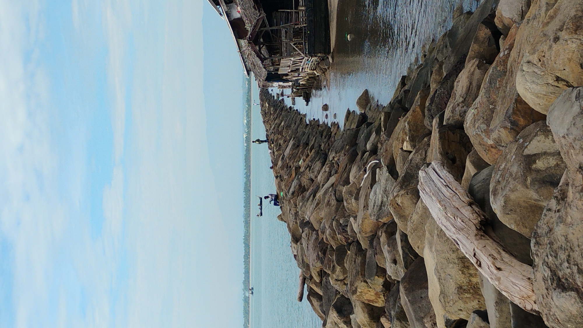 This is a vertical, eye-level, full-shot photograph of a rocky jetty extending into the ocean in Lhokseumawe, Indonesia. The jetty is constructed of large, rough-hewn stones. The sea is a calm, muted blue-green, with a gentle ripple on its surface. In the distance, the coastline is visible, marked by a line of palm trees and buildings. The sky is a pale blue with scattered white clouds.
On the jetty, several individuals are present. In the foreground, near the base of the jetty, a large piece of driftwood lies on the rocks. Further along the jetty, a man sits on the rocks, fishing. He is casting his line into the water. Another man stands further up the jetty, facing away from the camera. In the middle distance, a small boat with two people on board sails on the ocean.
To the right of the jetty, a rustic, wooden structure is built on stilts over the water. It has a thatched roof and appears to be a simple dwelling or shop. The structure is weathered and shows signs of disrepair. The water around the structure is calm and reflects the sky.
The overall impression is one of a tranquil, coastal scene, likely a fishing village or a recreational area along the coast of Lhokseumawe. The image captures a moment of everyday life in this Indonesian city.