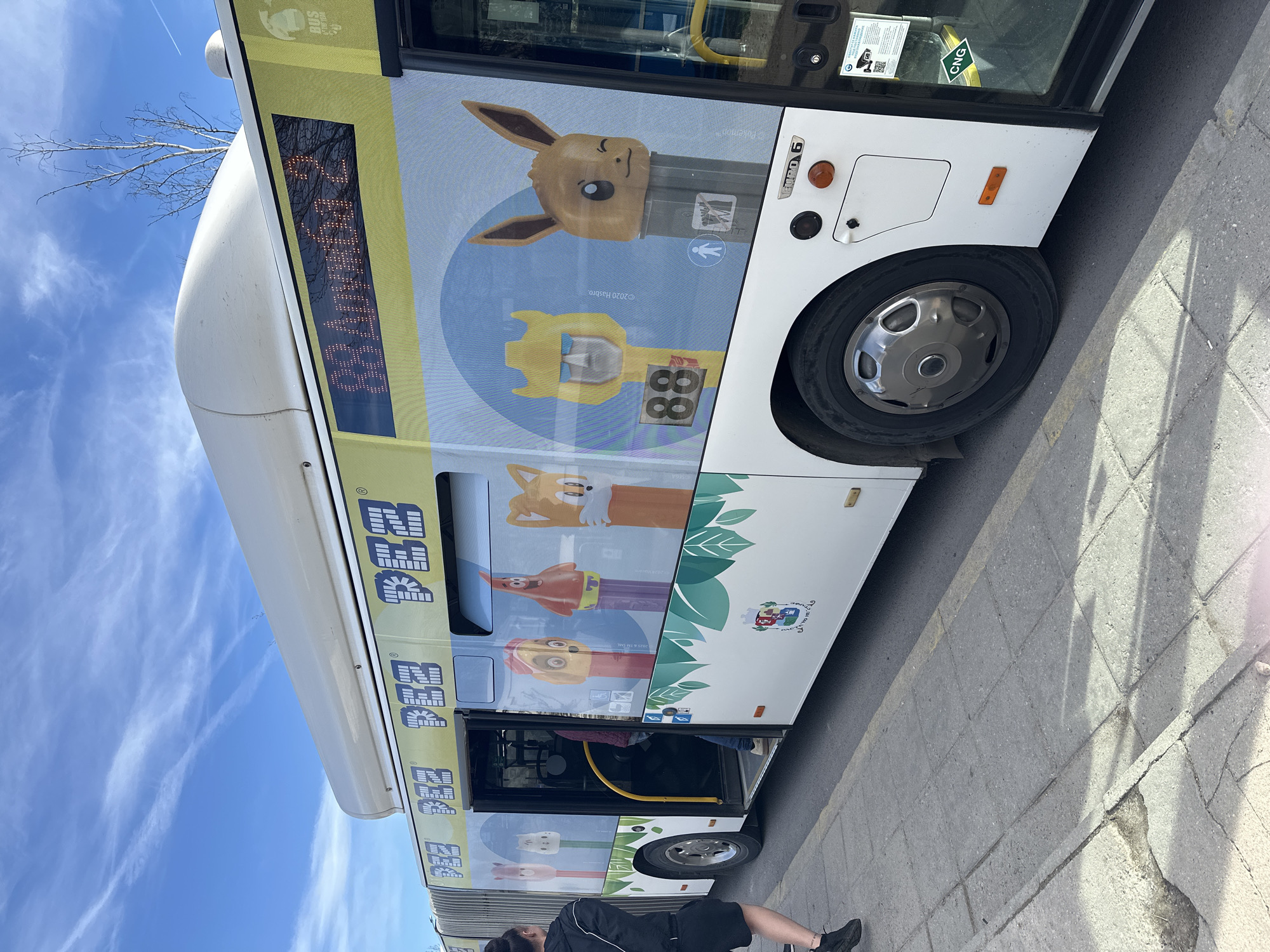 The image shows a bus with Pokémon-themed Pez dispenser designs on its windows, suggesting it is a promotional vehicle of some kind. The bus is white with a yellow stripe at the top, displaying the word "PEZ."
One window features an Eevee Pez dispenser. Another displays a fox, a yellow giraffe, and a reddish-orange character (possibly Patrick Star from Spongebob). Green leaf graphics adorn the bottom of the bus design. A logo with a castle and writing in Bulgarian is also present near the bottom.
The bus is parked alongside a grey stone sidewalk or road. A person is standing near the bus door.
The bus's electronic sign displays text (likely the bus route). The bus has a 'CNG' sticker indicating it runs on compressed natural gas. The image was likely taken outdoors on a sunny day. It is in Sofia, Bulgaria, indicated by the Bulgarian text on the bus's logo.