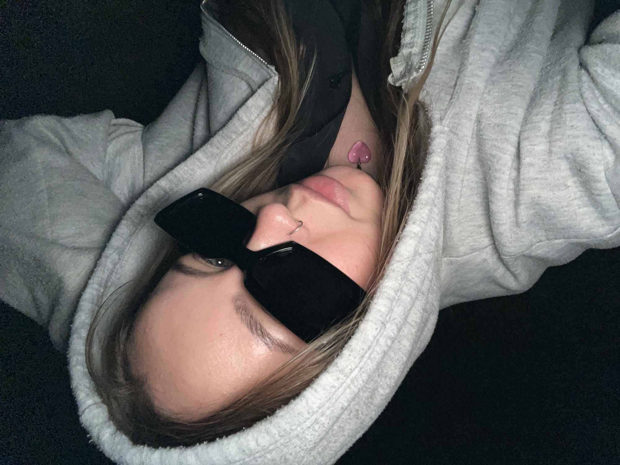 A selfie-style photograph presents a young woman with fair skin and light brown hair, wearing dark sunglasses and a light gray hooded sweatshirt. A black garment is visible beneath the sweatshirt. She has a nose ring in her left nostril and a pink heart-shaped object, possibly jewelry or a tattoo, is visible on her neck. The background is dark and indistinct. The image appears to be rotated counter-clockwise. There is no information in the image to suggest it was taken in Novopokrovskaya, Russia.