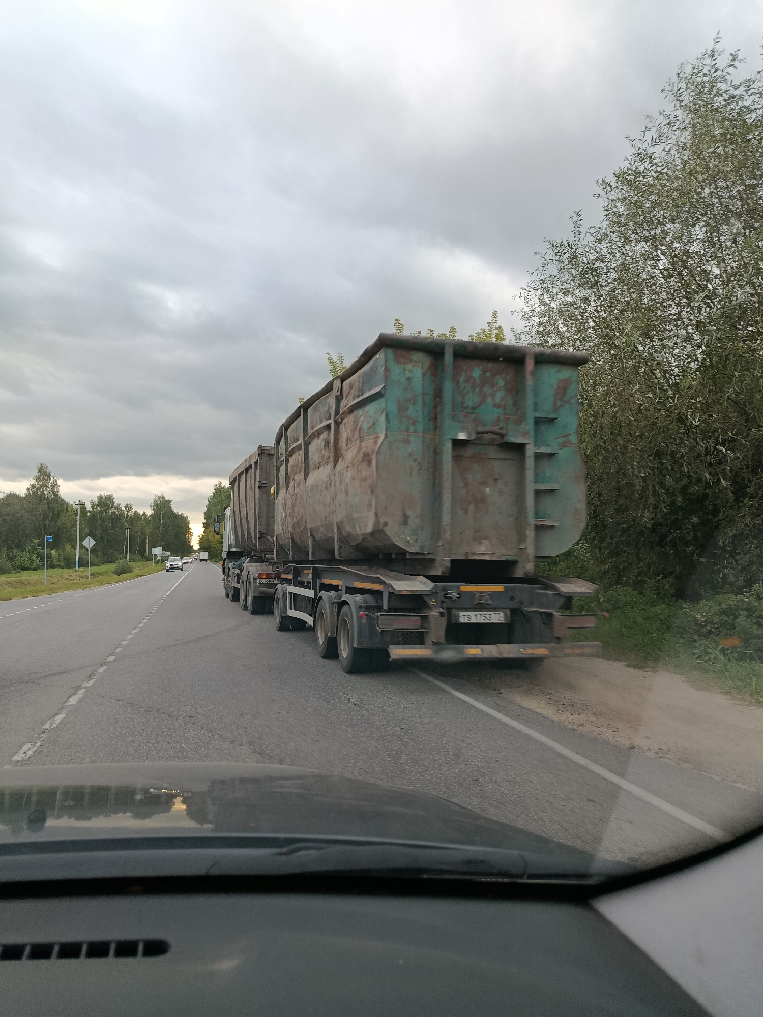 The image shows a truck with two large, green, and rusty dumpsters on a highway in Verereya, Russia. The truck is moving on the right lane of a two-lane road. The truck's license plate is partially visible and reads "T8 1753 72". The road is surrounded by trees and vegetation. The sky is overcast with clouds. The image is taken from inside a car, showing the dashboard and windshield in the foreground.