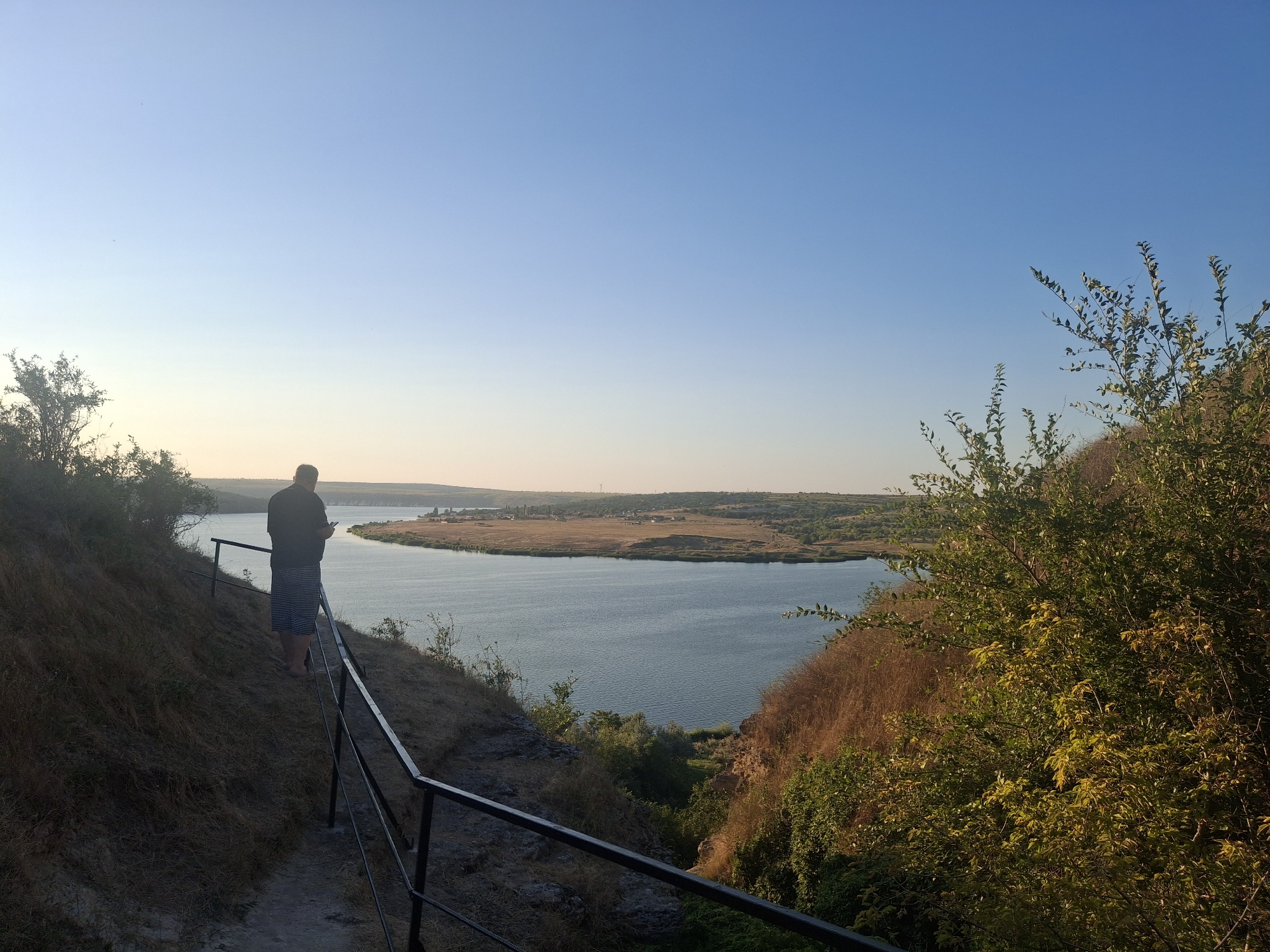 A man stands on a path overlooking a wide river in Roghi, Moldova. The man is facing away from the camera, looking out at the water and the distant shore, which is lined with dry, golden fields and sparse vegetation. He is wearing a dark t-shirt and striped shorts. A black metal railing runs along the edge of the path, which is made of dirt and gravel. The sky is clear and a light blue, with a soft glow indicating either early morning or late afternoon. The overall scene is peaceful and serene, with nature dominating the landscape.