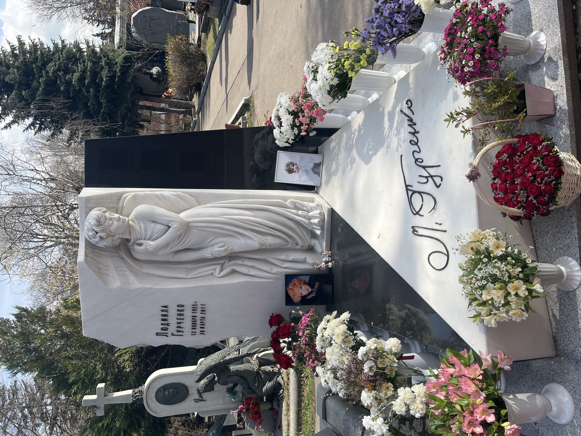 The image shows a grave in what appears to be a cemetery in Moscow, Russia. The large, elaborate headstone features a marble sculpture of a reclining angel. The headstone is inscribed with the name "Lyudmila Gurchenko" and dates: "12 ноября 1935 - 30 марта 2011" which translates to 12 November 1935 - 30 March 2011. Two framed photographs of Gurchenko are placed on the grave. Numerous bouquets of flowers, including roses, lilies, and other varieties, are arranged around the base of the headstone and in vases atop the marker. Other graves and trees are visible in the background, indicating a cemetery setting. A paved walkway is visible in the top of the image.