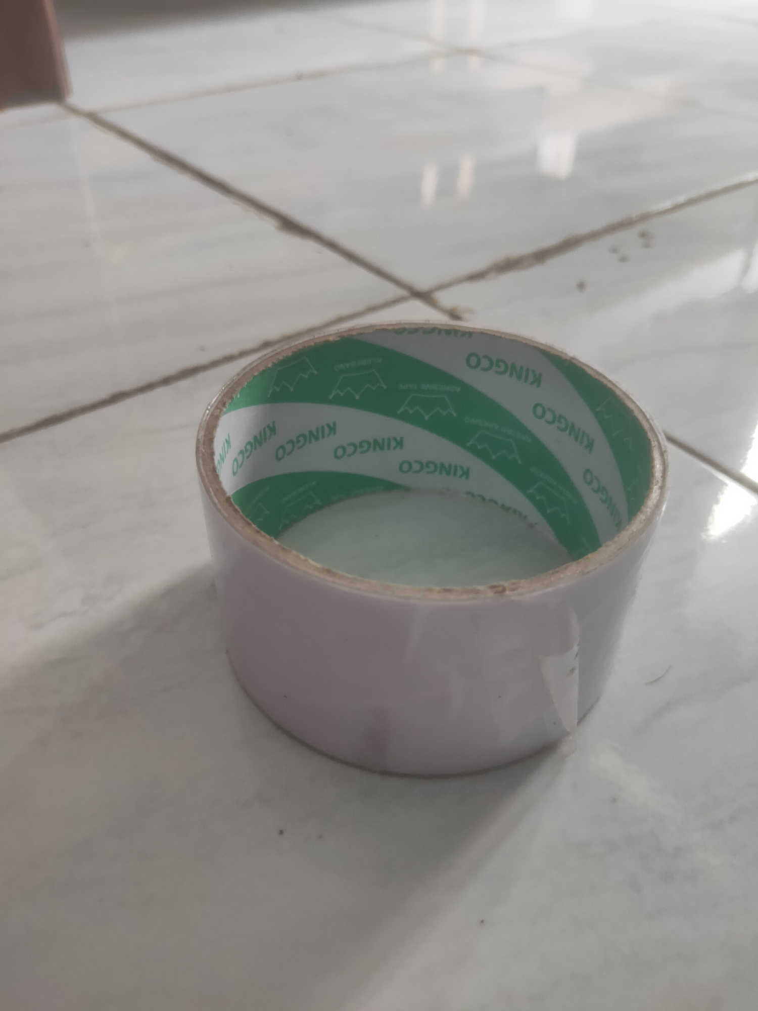 A roll of white double-sided tape with green branding is positioned on a tiled floor. The tape roll is the primary subject, centered in the frame and angled slightly towards the viewer. The green inner lining of the tape roll displays repeating text that appears to be "KINGCO". The surrounding environment is a floor composed of light-colored square tiles, with visible grout lines. The lighting suggests an indoor setting, potentially in a room in Antirogo, Indonesia, though this specific location detail is inferred and not visually confirmed by the image itself. There are no people or other objects prominently featured in the scene.