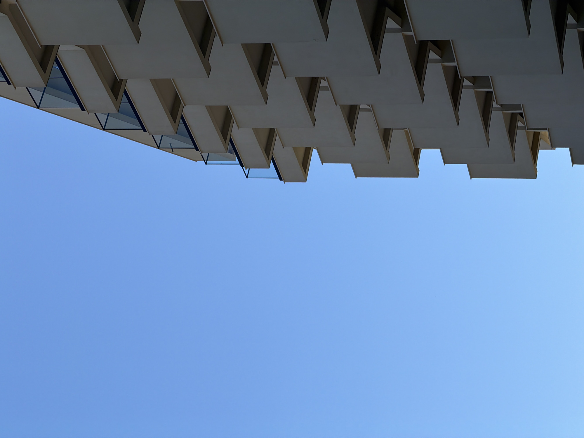 The image presents an upward view of a building's architectural detail against a clear blue sky. The building features a series of angular, repeating concrete or stone elements that create a saw-tooth pattern along its facade. Some sections reveal triangular glass panels behind the structural overhangs. The photograph is taken from a low angle, emphasizing the geometric design and its contrast with the unobstructed sky. The location is Boreti, Montenegro, and the structure is the Dukley Hotel, known for its distinctive facade.