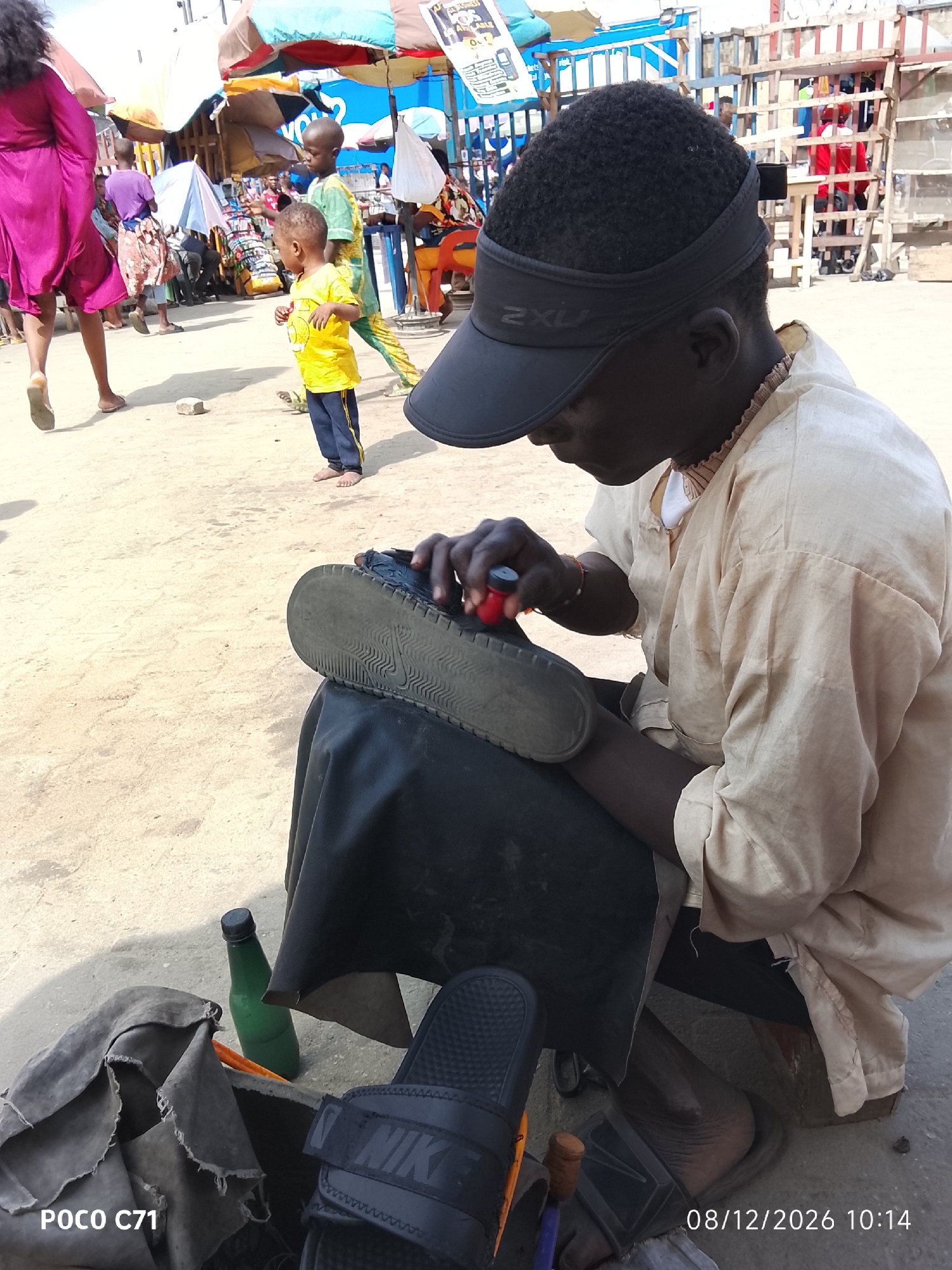 In Lekki, Nigeria, an outdoor market scene is depicted with several people present. In the foreground, a shoemaker is actively working on a shoe. He is wearing a light-colored traditional tunic and a black visor. His hands are engaged in polishing the sole of a black shoe, which rests on a black cloth. Adjacent to him, a green bottle and a pair of black Nike slides are visible. In the background, various market stalls with colorful umbrellas are set up. Children are also seen in the mid-ground; one boy in a yellow shirt and blue pants stands near a stall, and another child in a green and yellow outfit walks by. A woman in a vibrant pink dress is also in the background. The ground is sandy and appears to be unpaved. The overall impression is a bustling marketplace environment.