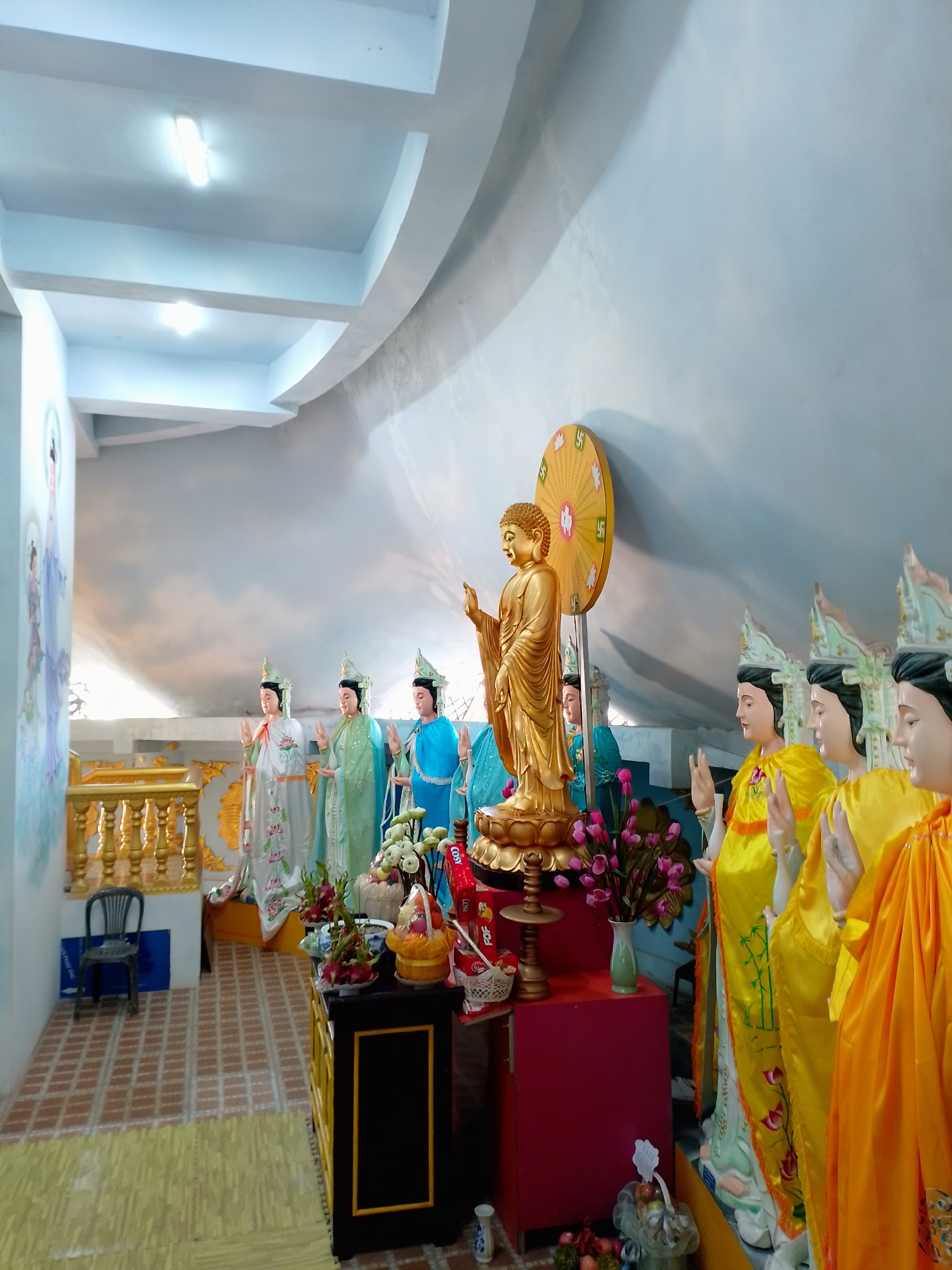 The image shows an interior scene in Thôn 3, Vietnam, featuring a Buddhist shrine with several statues. In the center, a golden Buddha statue stands on a lotus pedestal. Behind it is a large, circular, golden disc with swastika-like symbols. To the left of the Buddha are several statues of figures in robes of various colors, including white, green, and blue. To the right, there are statues with yellow robes. These figures are depicted with their hands raised in a gesture. In front of the statues, there are offerings of fruit and flowers on a black table with gold trim. The walls are pale and feature a painting on the left. The ceiling is multi-tiered with recessed lighting. The floor is tiled with a geometric pattern. A small blue chair is visible in the left background.