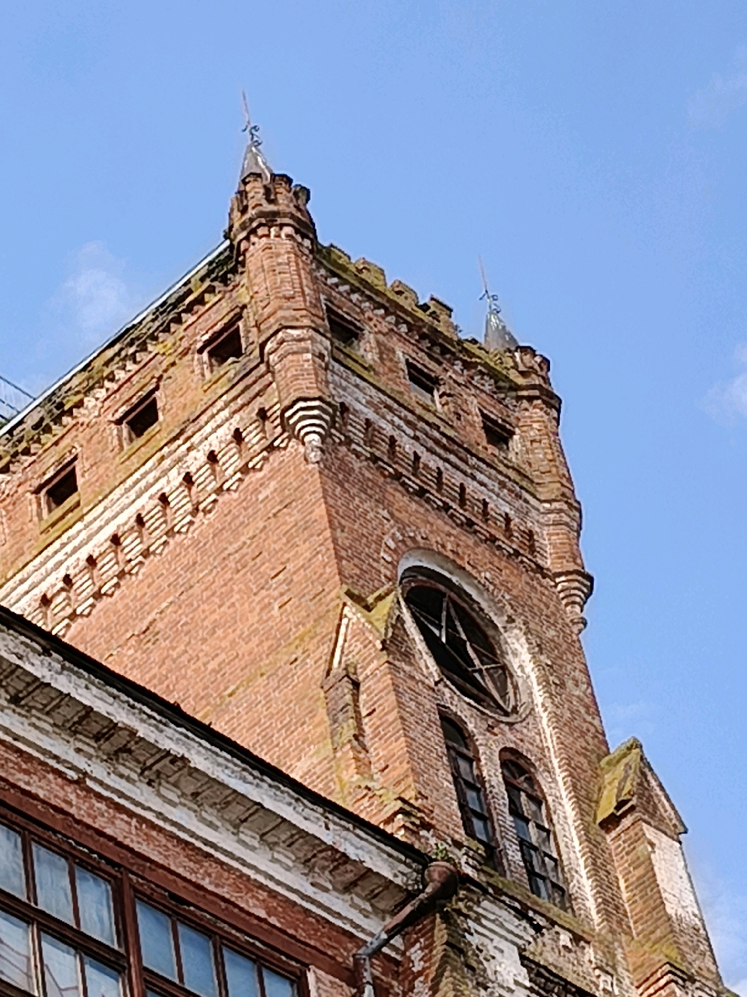 The image shows a close-up, low-angle view of an old, brick clock tower against a clear blue sky. The tower is made of red brick and features intricate detailing, including decorative brickwork around the windows and cornices. Several windows are visible, some appearing to be broken or boarded up. The tower has a pointed roof with spires. A section of an adjacent building with large windows is visible in the lower left corner. The scene is one of an old, possibly derelict, architectural structure. The specific location of this structure in Naro-Fominsk, Russia, is not discernible from the image itself. There are no people or animals present in the image.