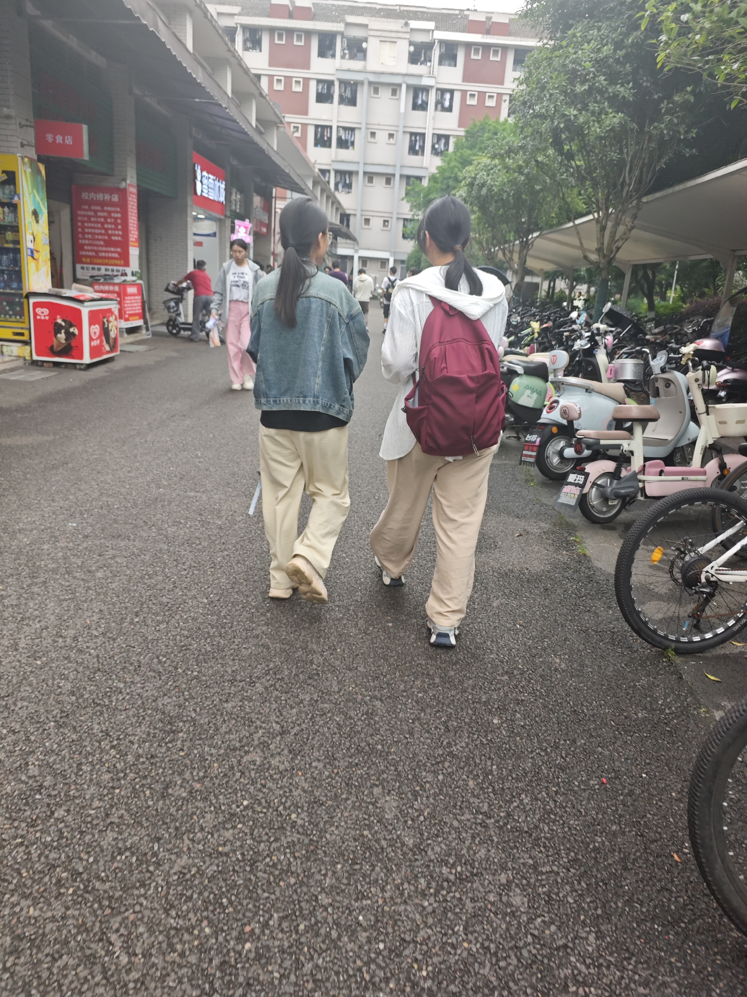 Two young women, one wearing a denim jacket and the other a white hooded jacket, walk away from the camera on a paved street in the Chongqing High-tech Zone, China. The woman on the left has her hair in a ponytail and is wearing loose-fitting light-colored pants and sneakers. The woman on the right carries a maroon backpack and is also wearing light-colored pants and sneakers. The street is lined with buildings and parked electric scooters and bicycles. A red and white ice cream display is visible on the left side of the frame, along with a vending machine. Trees and an overhang structure are also present in the background. The overall scene depicts a typical day in an urban environment.