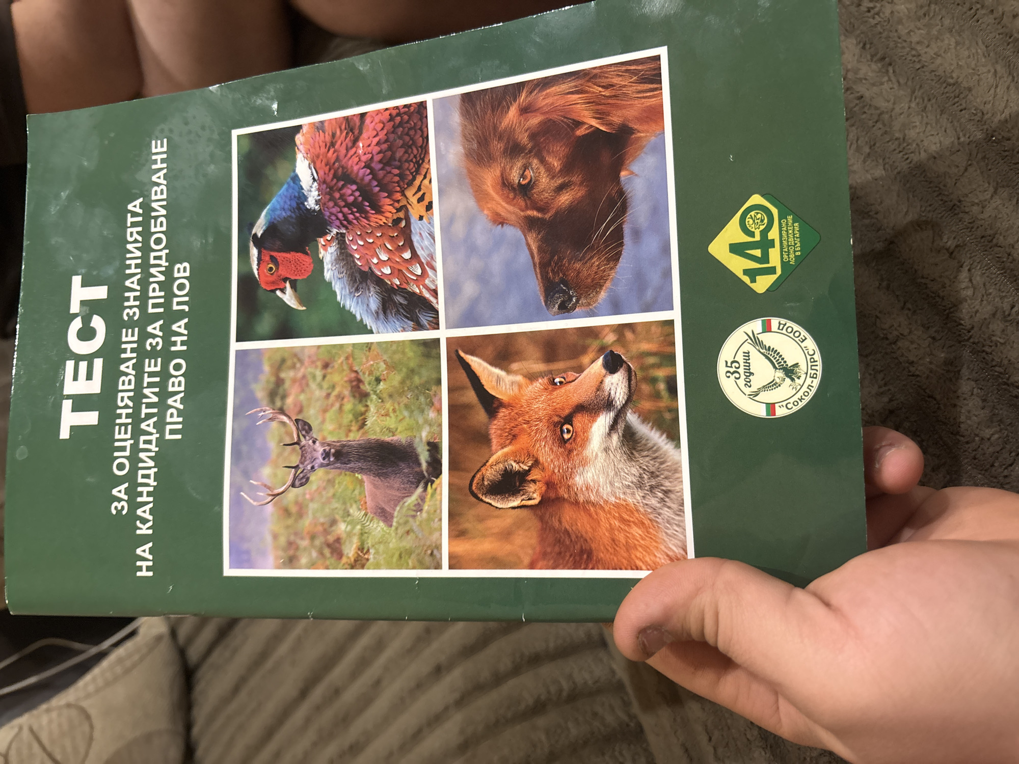 The image shows a book cover held by a person's hand. The book's title, in Bulgarian, translates to "Test for Assessing Knowledge of Candidates for Hunting Rights." The cover features a green background with four animal images arranged in a grid: a pheasant, a dog, a deer, and a fox. Two logos are visible on the right side of the cover. The background appears to be a brown textured surface. The photo seems to be taken indoors. The book is likely about hunting regulations and knowledge required for obtaining hunting rights in Bulgaria. There is no information available in the image concerning the city of Burgas, Bulgaria.