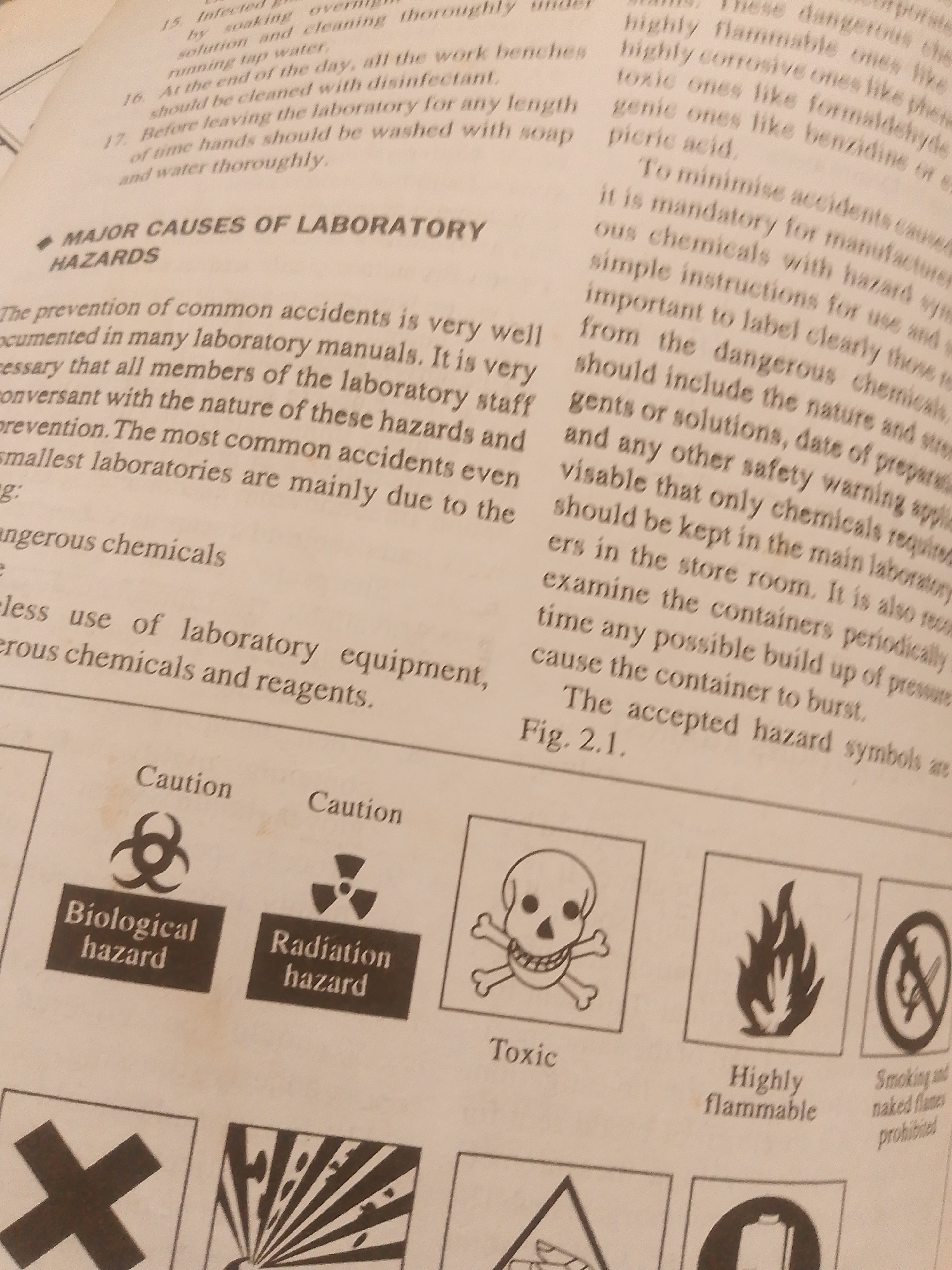 This is a photograph of an open book displaying text and hazard symbols. The book appears to be a manual or educational material related to laboratory safety.
The text discusses the prevention of common accidents in laboratories, emphasizing the importance of understanding hazards and following instructions. It also mentions keeping chemicals in the store room and checking containers for pressure buildup. The text is presented in two columns on the right side of the image and in paragraphs on the left.
Below the text, a grid of hazard symbols is visible. These symbols represent different types of dangers encountered in a laboratory. Visible symbols include:
* A biohazard symbol with the text "Biological hazard"
* A radiation symbol with the text "Radiation hazard"
* A skull and crossbones symbol with the text "Toxic"
* A flame symbol with the text "Highly flammable"
* A prohibition symbol over a flame with the text "Smoking and naked flames prohibited"
* Other symbols, partially visible, include an "X" shape and an exploding object.
The overall setting is indoors, likely in a room where such safety information would be relevant, such as a laboratory or a classroom. The lighting suggests an indoor environment. There are no people or explicit references to Jauro Yinu, Nigeria in the visible content of the image. The focus is on the educational content within the book.