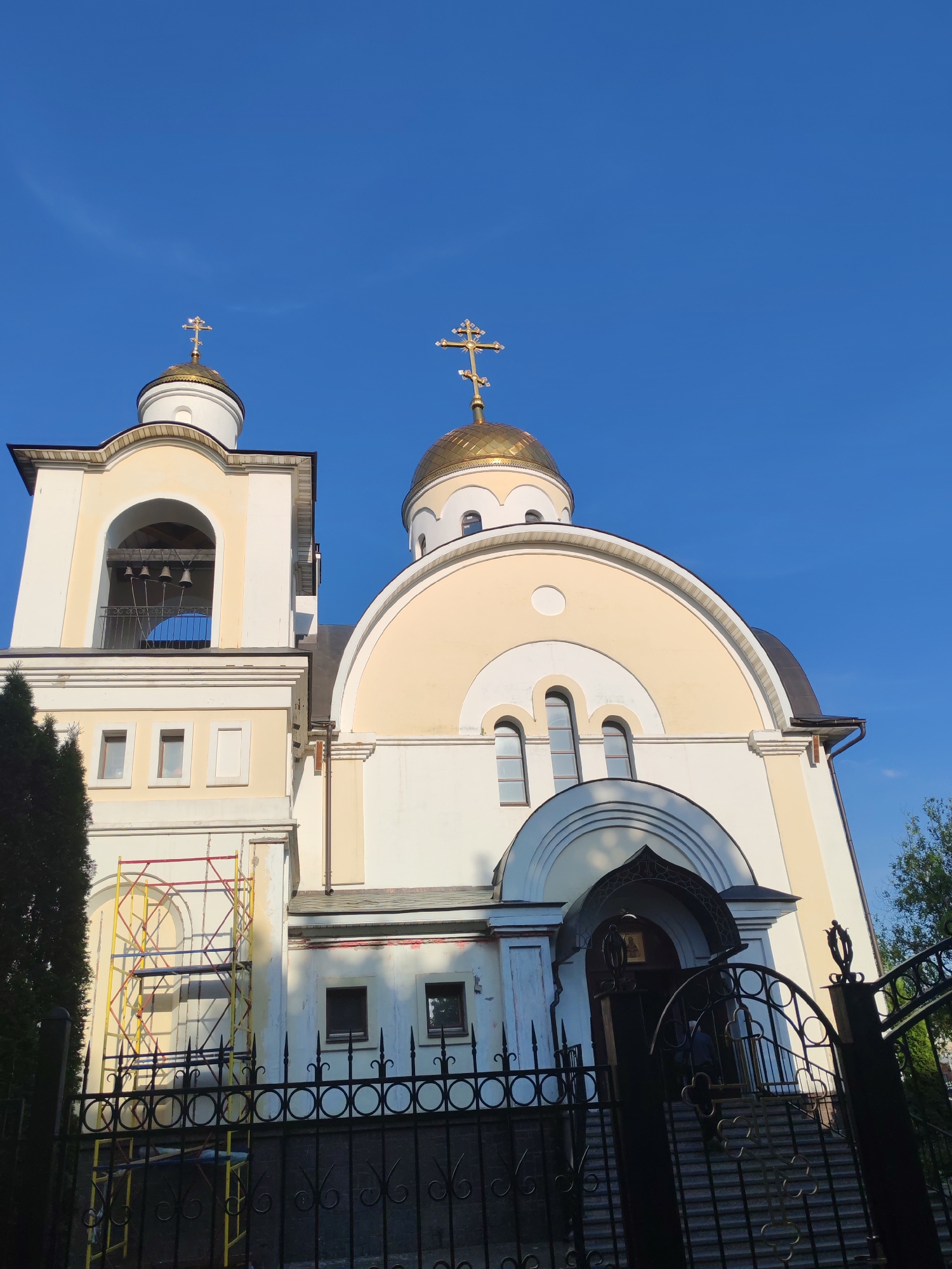 A low-angle shot reveals an Eastern Orthodox church with a cream-colored facade, under a clear, bright blue sky. The church features two domes, one gold and one lighter, each topped with a cross. A bell tower stands to the left, showing hanging bells within. Scaffolding is visible to the left of the tower. A black wrought iron fence runs along the front of the property, partially obscuring the building's entrance. The entrance, framed by an arched portico, leads to steps. The location is Podolsk, Russia.