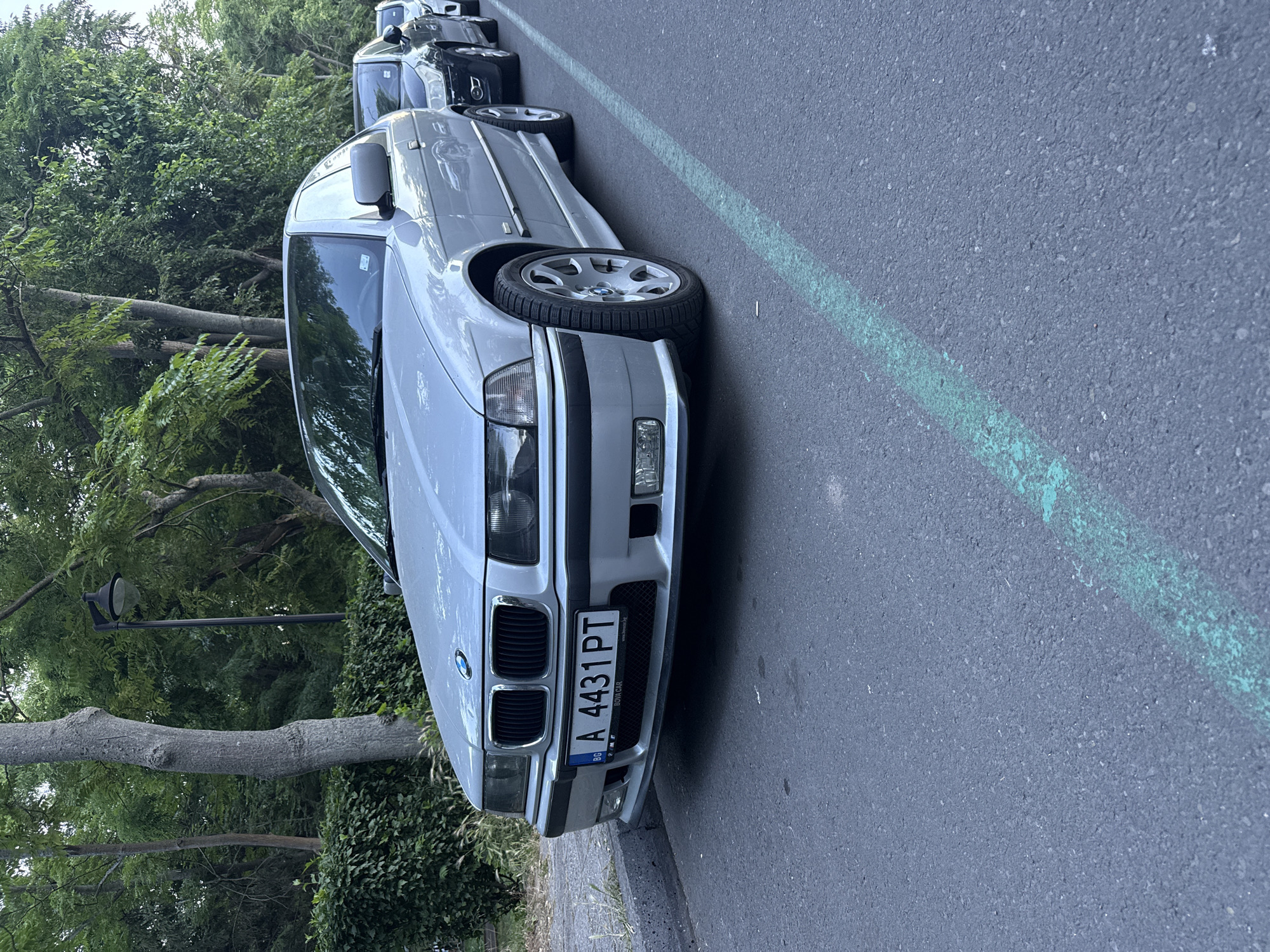 A silver BMW sedan is parked on the side of a paved road in Burgas, Bulgaria. The car has a Bulgarian license plate that reads "A 4431 PT". The car is facing towards the right of the frame, and another car, a dark sedan, is parked behind it. The road has a green painted line running along it, marking a parking space. In the background, there are trees and bushes, with a street lamp visible among the foliage. The scene appears to be in a residential or park area of Burgas.