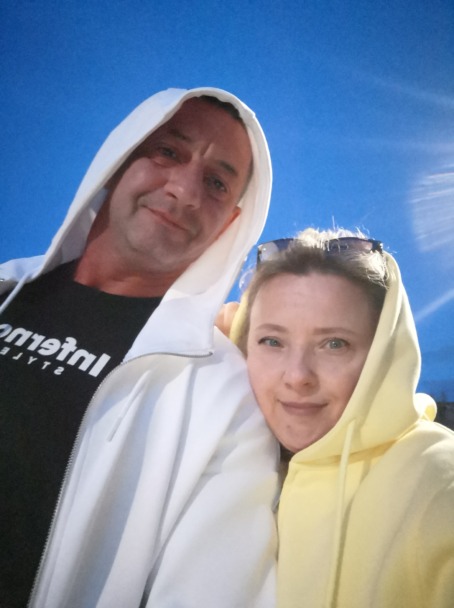 In an outdoor, urban setting, two individuals, a man and a woman, pose for a selfie. The man, with short hair and a light complexion, wears a white hooded jacket over a black t-shirt with white text. The woman, also with light skin and blonde hair, sports a yellow hooded sweatshirt and sunglasses perched on her head. They are positioned in the foreground, close to the camera, with a clear blue sky filling the background. The overall scene suggests a casual moment, possibly in the city of Alexandrovsk, Russia.