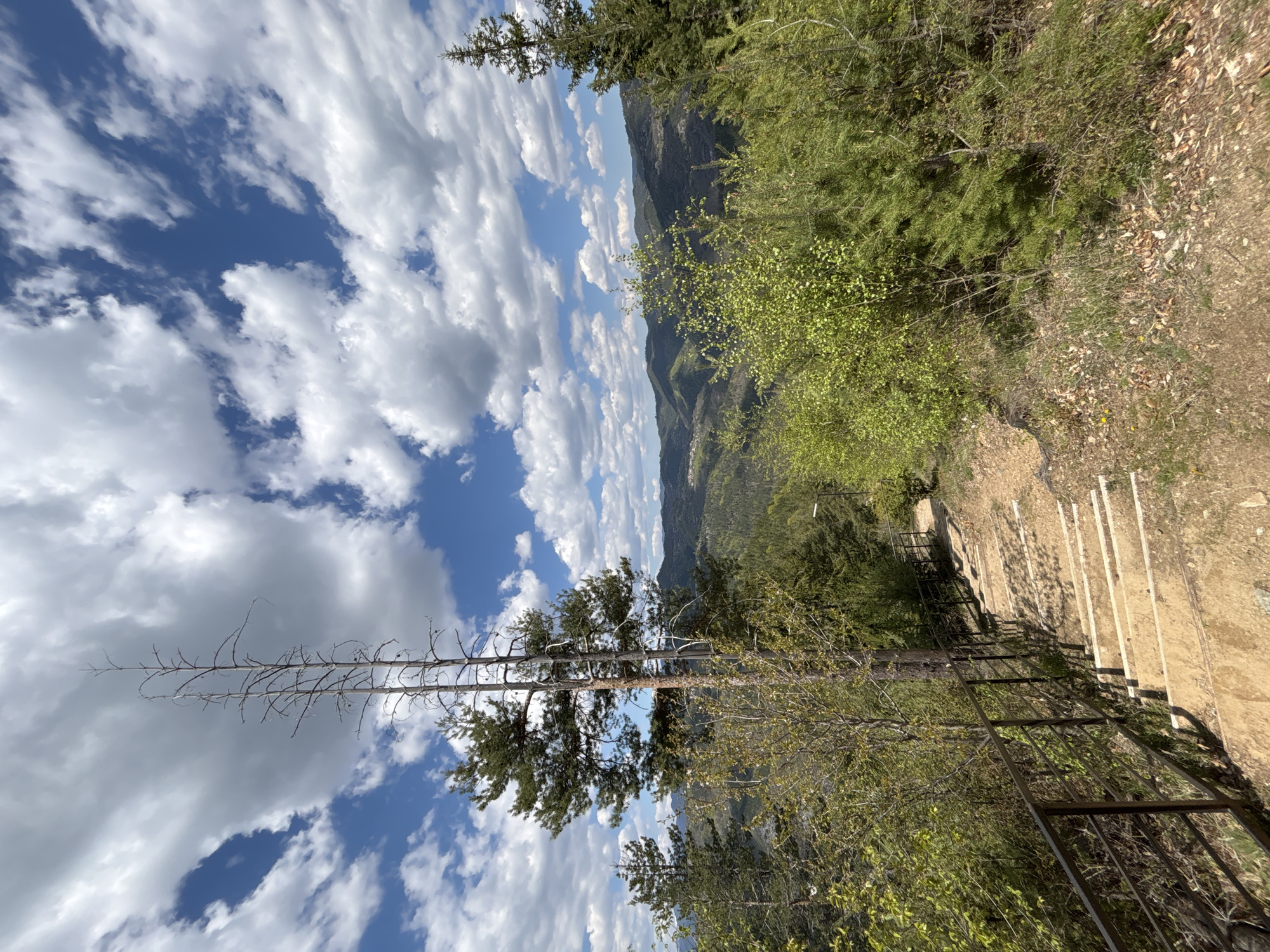 The image shows a mountain vista with a cloudy sky. The scene is captured from a high vantage point, possibly a trail or overlook. The right side of the image shows a descending path with wooden supports and a metal railing. Vegetation includes trees, shrubs, and ground cover. In the distance, mountain ranges are visible. The sky is blue with white, puffy clouds. The image is likely taken near Gradat, Bulgaria.
