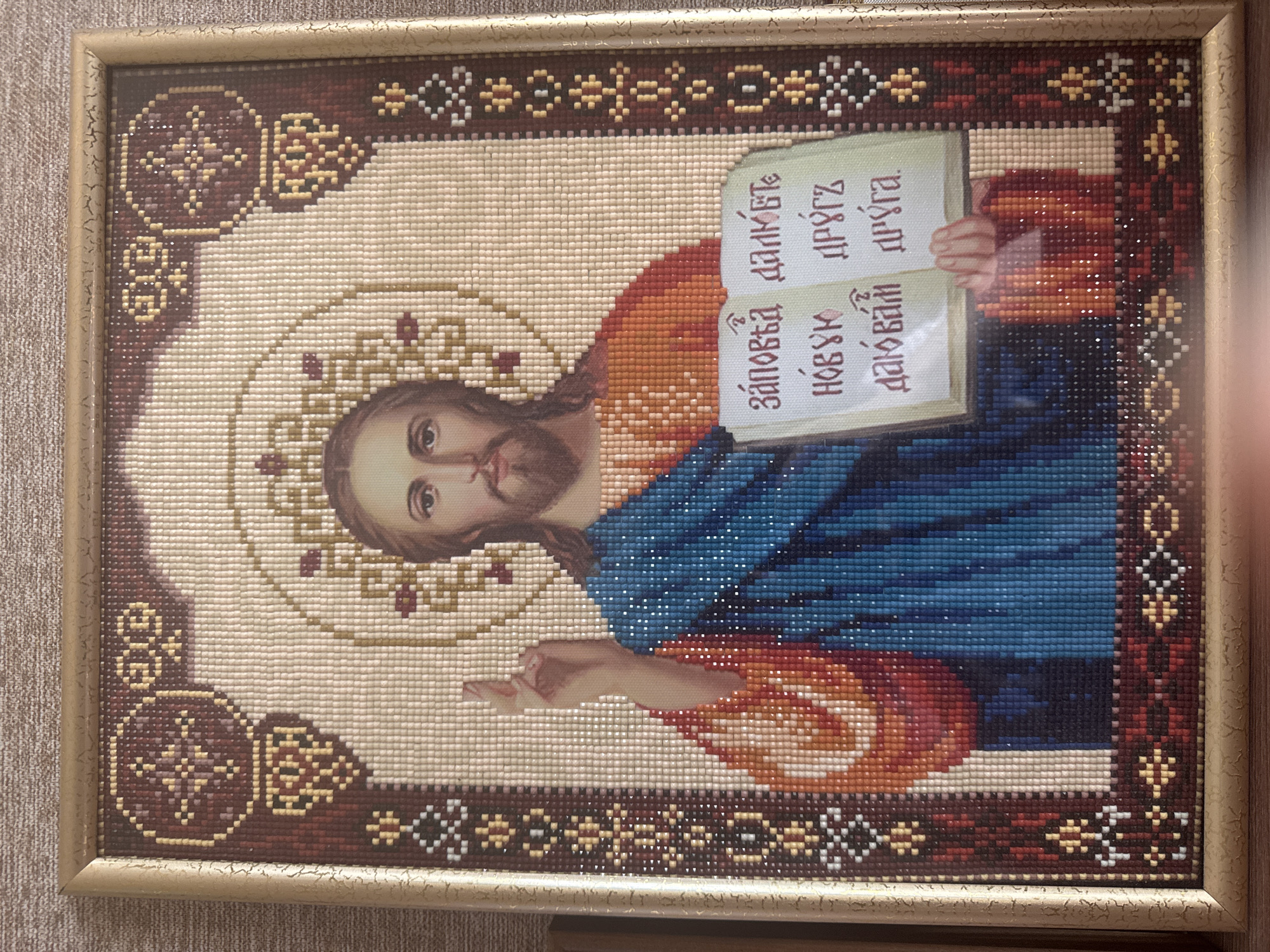 This is a framed diamond painting depicting Jesus Christ. Jesus is shown from the chest up, with his right hand raised in a gesture of blessing. He has a beard and is wearing a blue robe over an orange garment. He is holding an open book with text written in Cyrillic script. The background is a cream color, with an ornamental border in shades of brown, orange, and yellow. The frame is a light gold color. The diamond painting is likely a religious icon or artwork. There is no information about its location in the city of Яглово, Russia.
