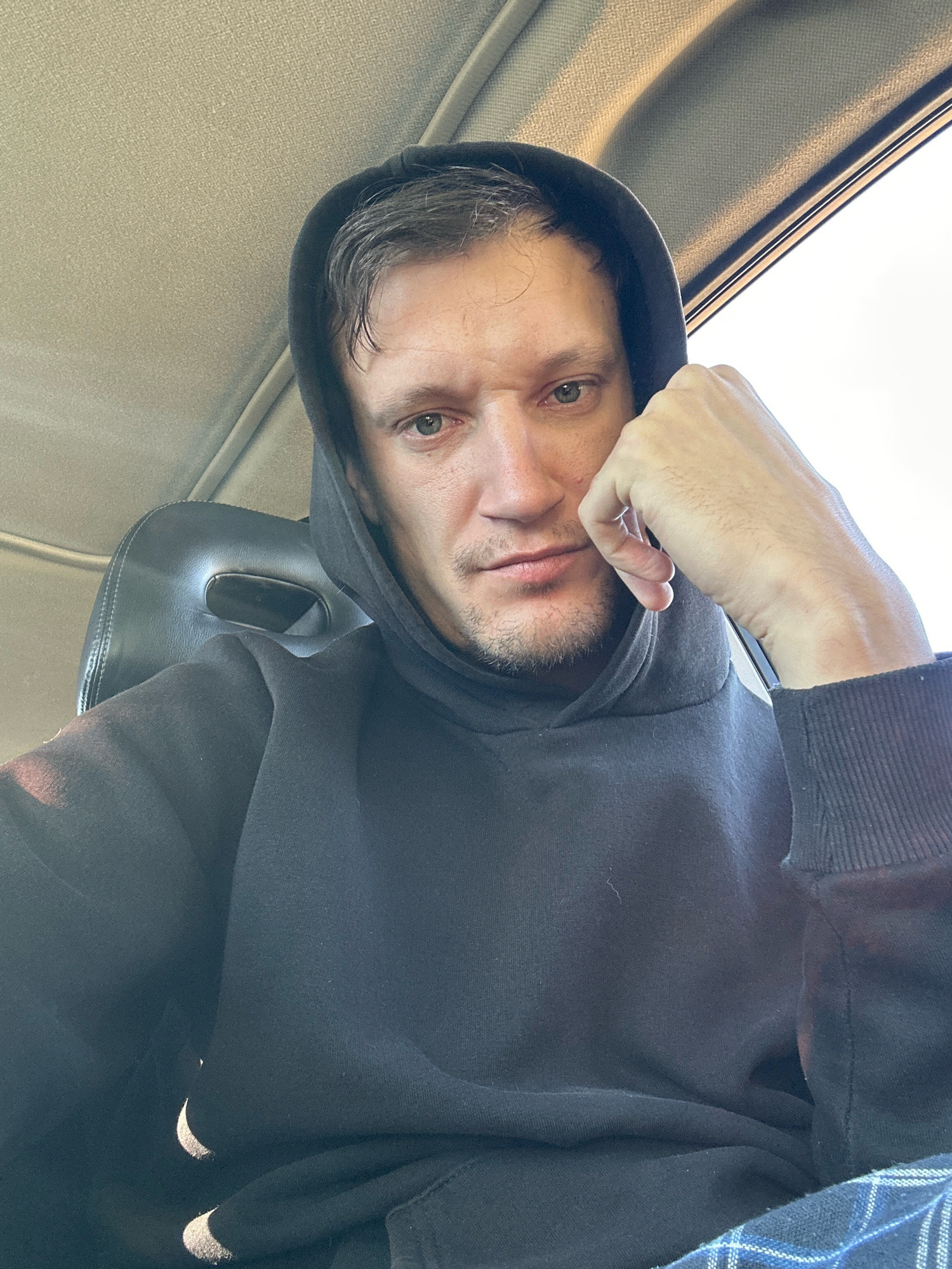 A Caucasian man with short, dark hair and light-colored eyes is pictured in a close-up, head-and-shoulders shot. He is wearing a black hooded sweatshirt. His right hand is resting on his chin, with his fingers partially covering his mouth. The man appears to be indoors, possibly in a vehicle, given the presence of a car seat visible behind him and the textured ceiling. The lighting suggests it is daytime. The background includes a window with a bright exterior view and the interior of a car. The scene is not definitively located in Makiv, Ukraine.