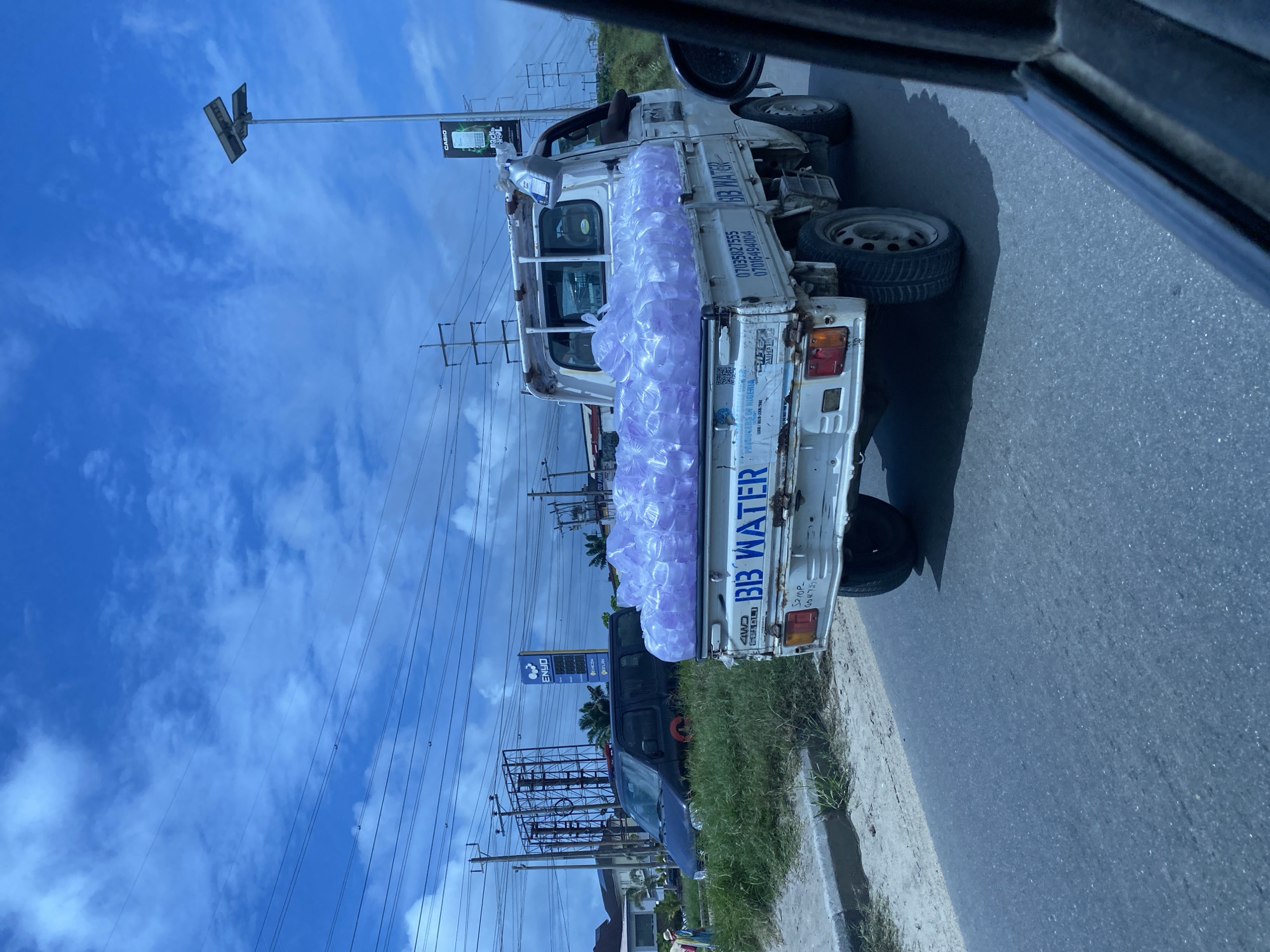In Lekki Peninsula II, Nigeria, a white pickup truck is loaded with numerous plastic bottles filled with water, possibly for delivery. The truck is driving on a paved road under a bright, cloudy blue sky, with power lines and utility poles visible in the background. A dark-colored van is parked on the grassy shoulder of the road. The scene appears to be in a semi-urban or commercial area, suggested by the presence of the gas station signage and advertising billboards. The overall atmosphere is casual, with the focus on the transportation of goods.