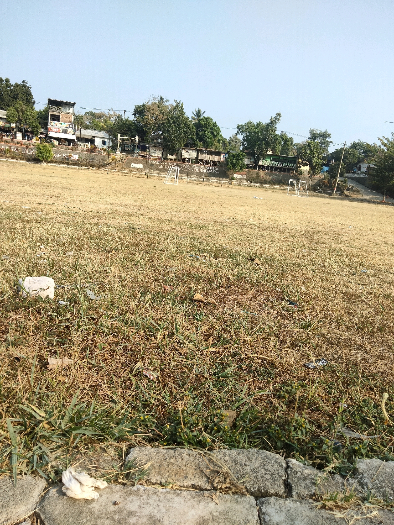 The image depicts an outdoor scene, likely a sports field, under a clear sky. The foreground is dominated by dry, sparse grass and some patches of greener growth, with a few pieces of white debris and some rocks. In the mid-ground, the field appears to be made of dirt or dry grass, with a white soccer goal visible. The background features a row of buildings, some with multiple stories, set against a backdrop of trees. The buildings are clustered together and appear to house businesses or residences. The overall impression is of an undeveloped or semi-developed area in a town or city. Based on the provided information, this scene is located in Sawahan, Indonesia.