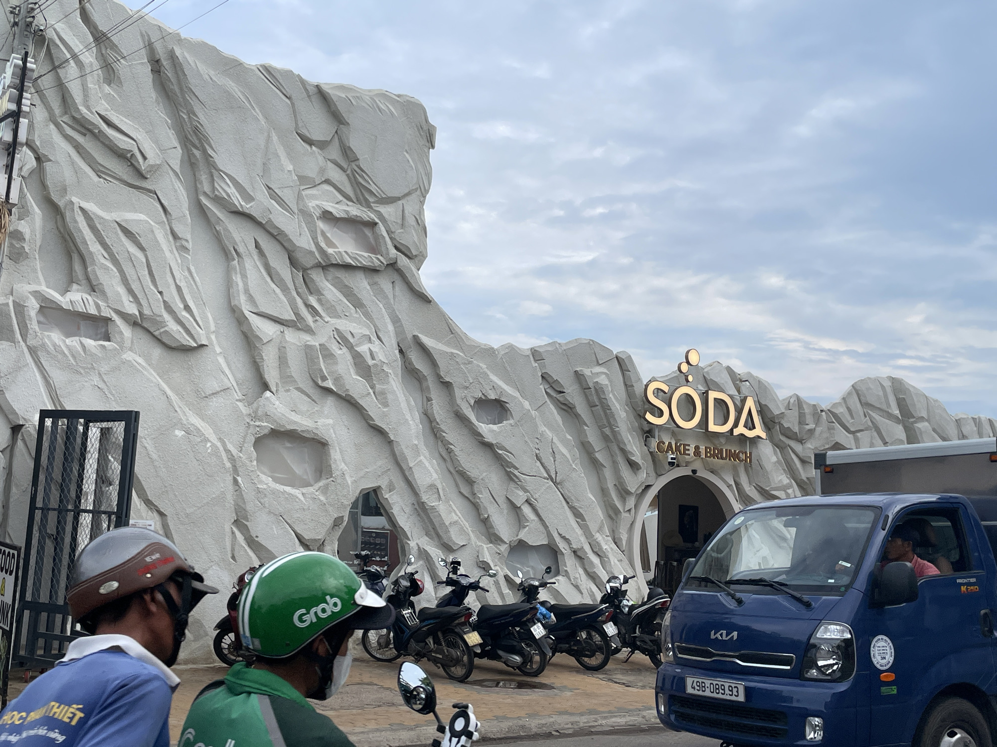 The image shows the exterior of "Soda Cake & Brunch" restaurant located in Phu Thuy Ward, Vietnam. The building has a distinctive facade designed to resemble a rocky cliff, with cave-like openings. Several parked motorcycles are visible in front of the restaurant. A blue KIA Frontier K250 truck is in the foreground, partially obscuring the view of the restaurant. Two individuals on a motorbike are positioned on the left side of the frame; one wears a blue shirt, while the other is wearing a "Grab" green jacket and a green helmet, suggesting they are a delivery driver. The sky is overcast.
