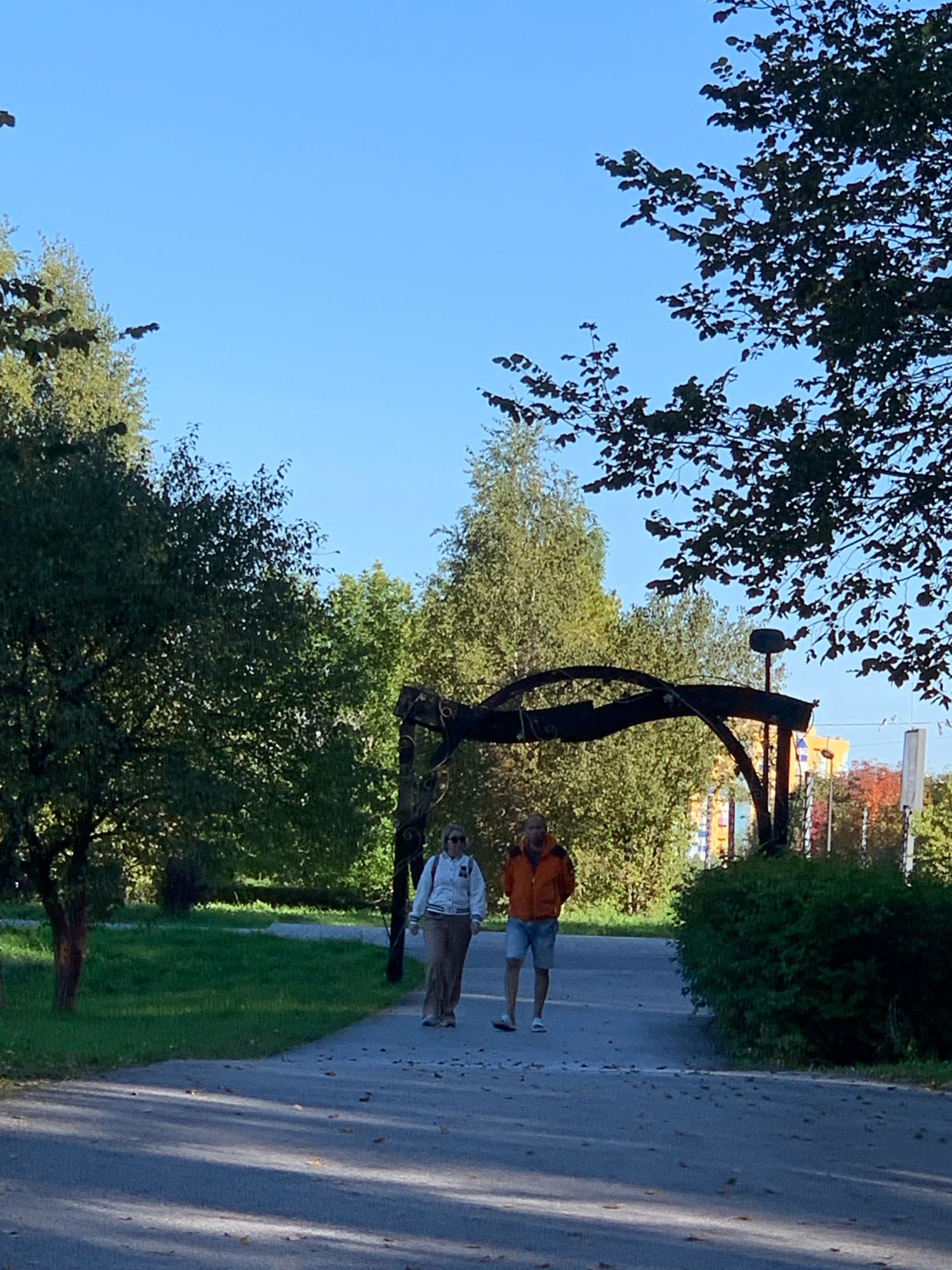 In Prokopyevsk, Russia, two individuals are walking on a paved pathway through a park on a clear day. The man, wearing an orange jacket and blue shorts, is to the right of the woman, who is in a white jacket and brown pants. They are walking away from the camera, towards a large, arched metal sculpture. Trees with green foliage are visible on either side of the path, and the ground is littered with fallen leaves. In the background, there are buildings and more trees. The scene appears to be set in a public outdoor space during autumn.