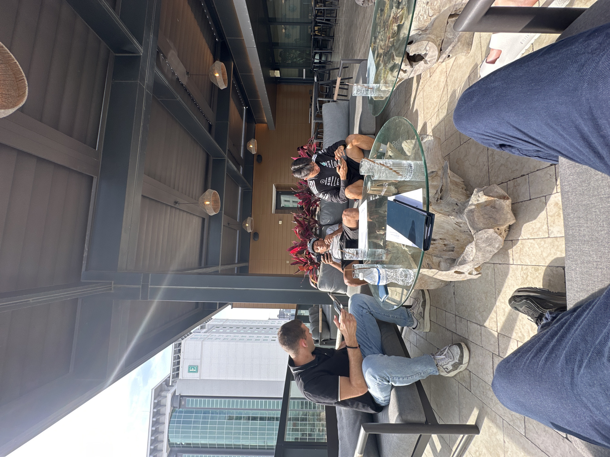 The image captures an outdoor gathering on a rooftop patio in Tampa, Florida, United States. Three individuals are seated around glass-topped tables with unique driftwood bases. The setting includes cushioned seating in a neutral color palette, set against the backdrop of the city skyline, seen through a glass barrier. The architecture includes modern high-rise buildings and industrial-style overhead structural elements, some supporting woven light fixtures.
One individual, wearing a black polo shirt and light-wash jeans, is looking at his phone. Another person is wearing athletic gear and appears to be occupied with a phone. The third person is only partially visible with legs wearing blue jeans. Glasses and bottled water are visible on the table.