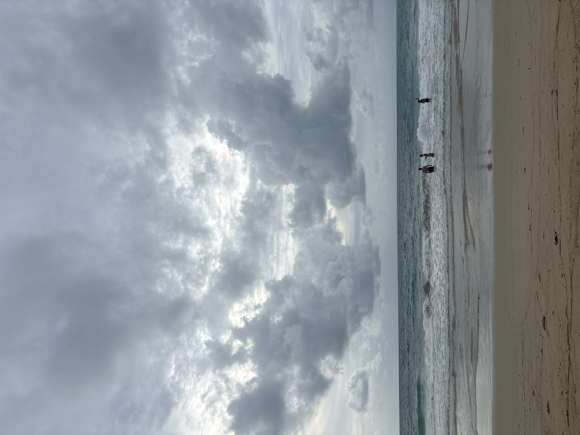 The image depicts a beach scene in Choeng Thale, Thailand under a heavily clouded sky. The beach is light tan, with wet sand reflecting the sky near the waterline. The sea is turbulent with visible waves. Three people are standing in the water at varying depths. One person is further out, partially obscured by a wave. The clouds are dense and grey, covering the majority of the sky.