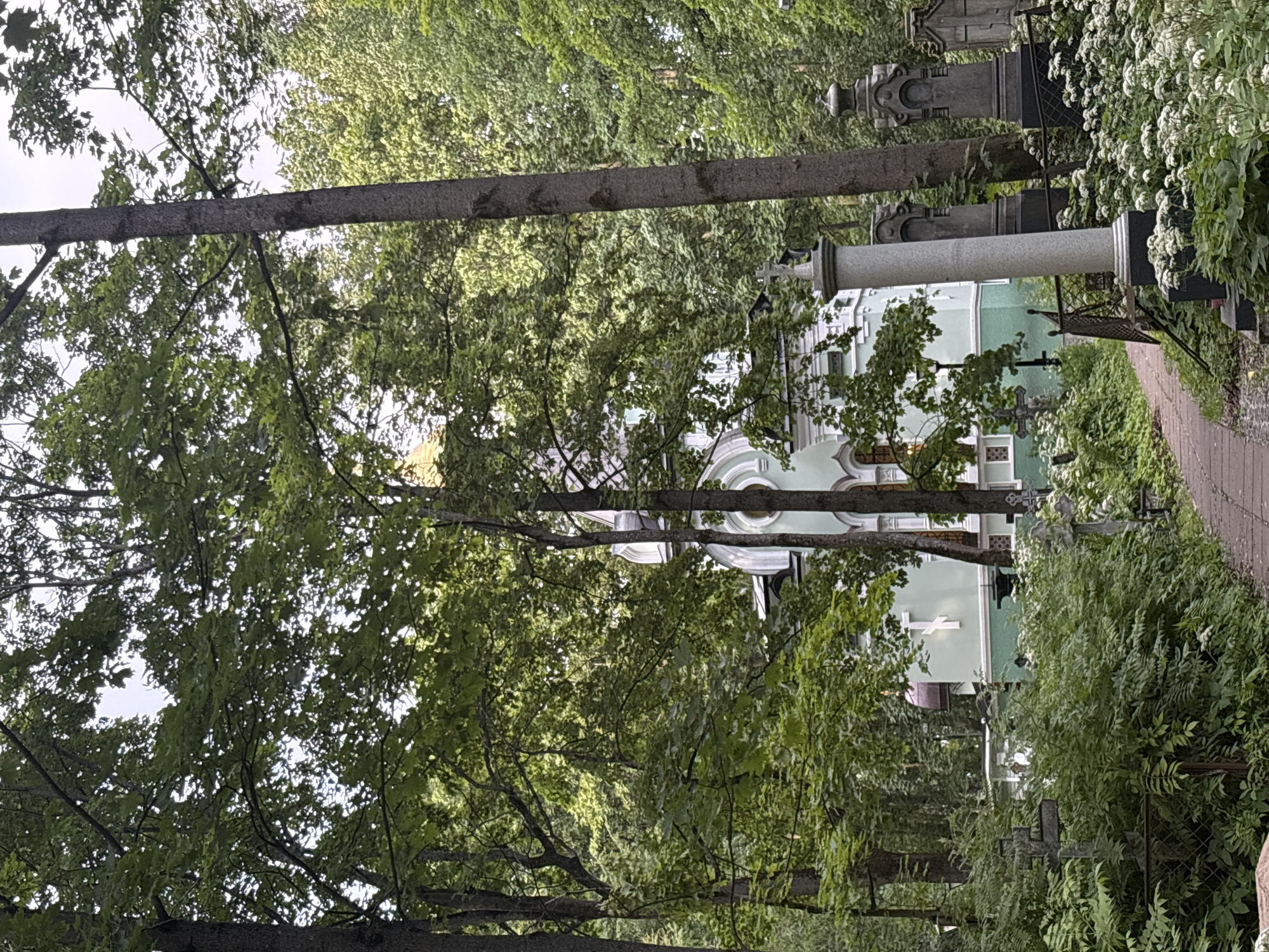 This media file depicts a cemetery in Saint Petersburg, Russia, during the daytime, likely on an overcast day given the soft lighting. The scene is dominated by lush green foliage from mature trees and dense bushes, which partially obscure the view of the cemetery structures.
Prominently featured are several gravestones and monuments, ranging from ornate, classical-style structures with columns and decorative elements to simpler crosses. A light green building, possibly a chapel or mausoleum, with white architectural details and arched windows is visible in the background. A large, light-colored column stands near the foreground, suggesting a significant monument. A paved path, appearing to be made of brick or stone, leads into the cemetery, bordered by a wrought-iron fence and abundant greenery.
The overall atmosphere is serene and solemn, characteristic of a cemetery. The dense trees and undergrowth suggest a well-established and possibly older cemetery. No people are visible in the frame, contributing to the quiet, contemplative mood. There is no visible text in the image.
