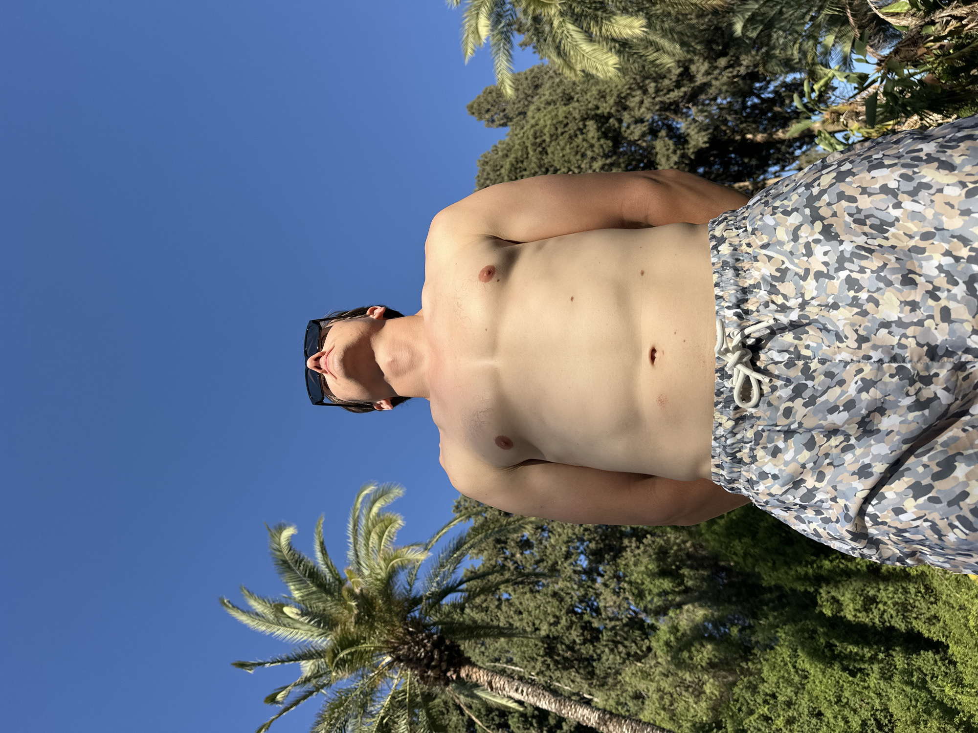 A man is standing outdoors, viewed from a low angle, looking up at a clear blue sky. He is shirtless, wearing patterned swim trunks with a drawstring. Sunglasses are on his face. Behind him, palm trees and dense green foliage are visible, suggesting a location with a warm climate. The overall impression is a sunny, relaxed, outdoor setting, possibly a garden or park in a place like Savyon, Israel, known for its greenery and pleasant weather.
