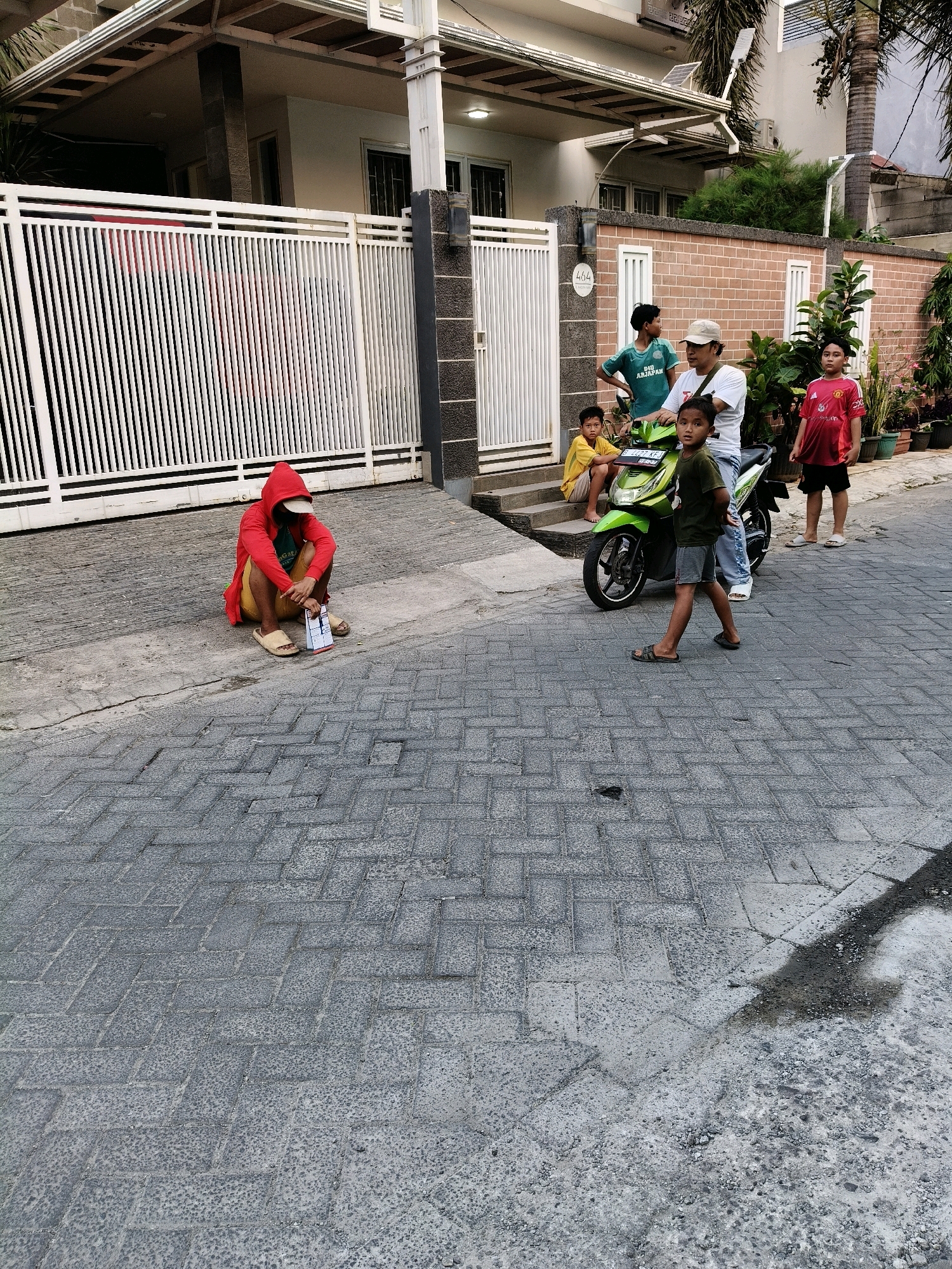 In the city of Bekasi, Indonesia, a scene unfolds on a paved street in front of a modern house. The house features a white metal fence and a red brick wall. A group of children are gathered. One child, wearing a red hooded jacket and yellow shorts, sits on the ground near the fence. Several other children and an adult are near a green motorcycle. One boy in a green shirt stands to the left of the motorcycle, while another boy in a red shirt stands to the right. An adult wearing a white shirt and a cap is seated on the motorcycle. A young boy in a dark green shirt and grey shorts stands in the foreground, facing the camera. The street is paved with interlocking grey bricks.