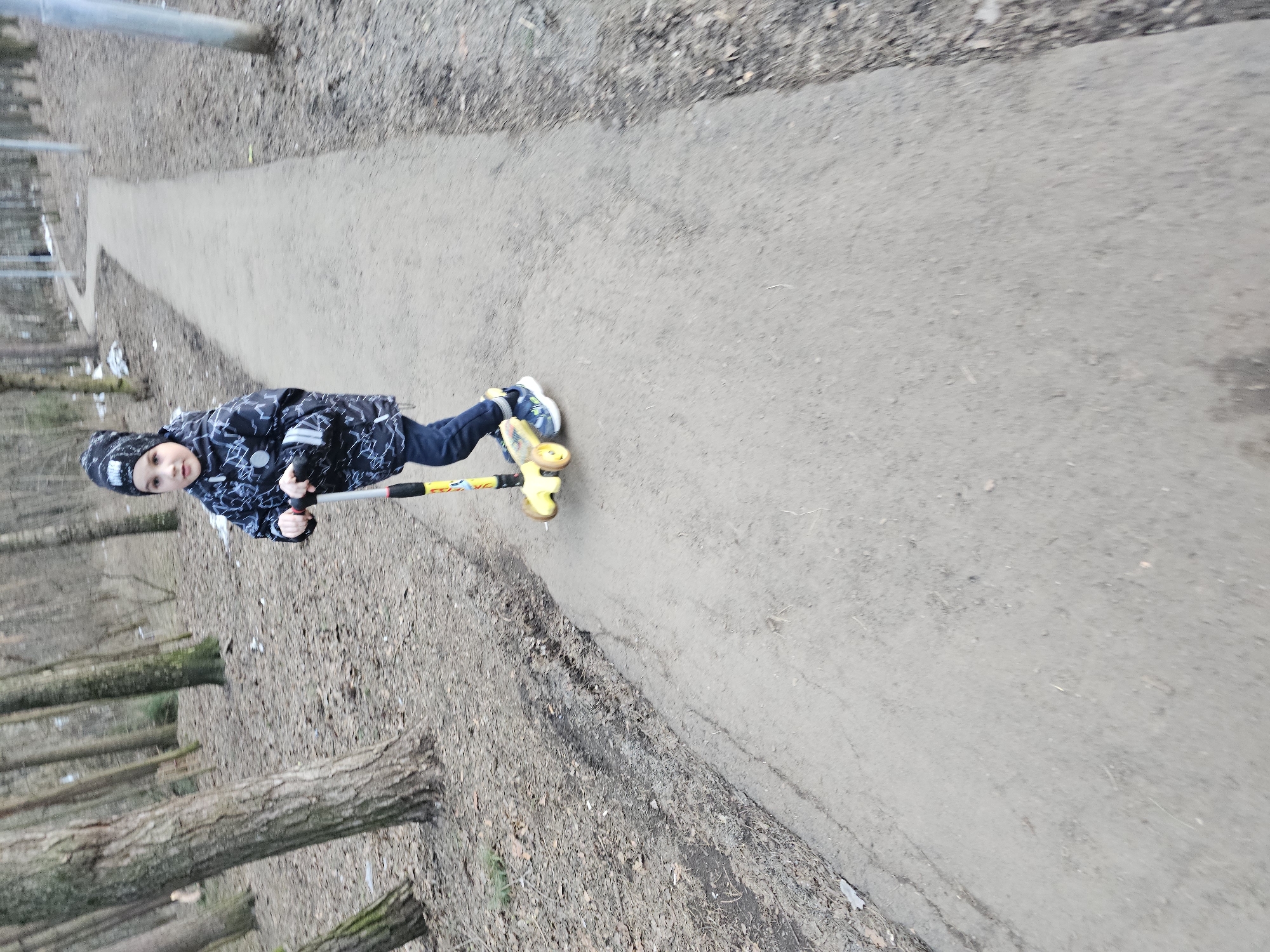 The image shows a young child riding a yellow scooter on a paved path. The child is wearing a dark patterned jacket and dark pants. The path is surrounded by trees and fallen leaves. The location is the city of Khimki, Russia.