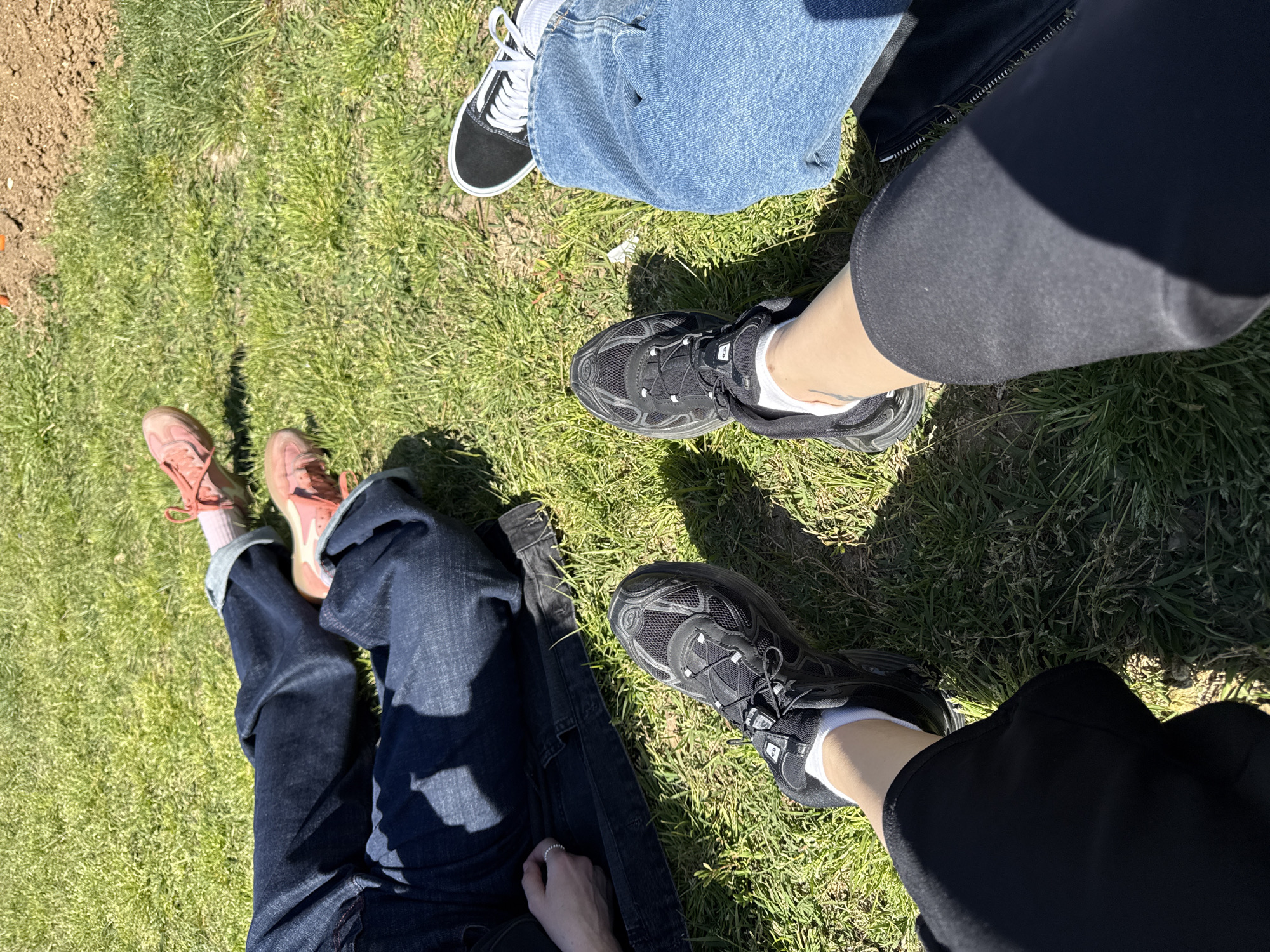 The image depicts a grassy area, possibly a park, with three individuals sitting or reclining. Two of the people are wearing black Salomon sneakers, with black leggings or trousers on one and jeans on the other, and white socks. The third individual is wearing pink sneakers with dark blue jeans. One person is wearing black Vans sneakers with blue jeans. All individuals appear to be relaxing outdoors on a sunny day. There is no indication of Barcelona, Spain, or any specific urban setting.