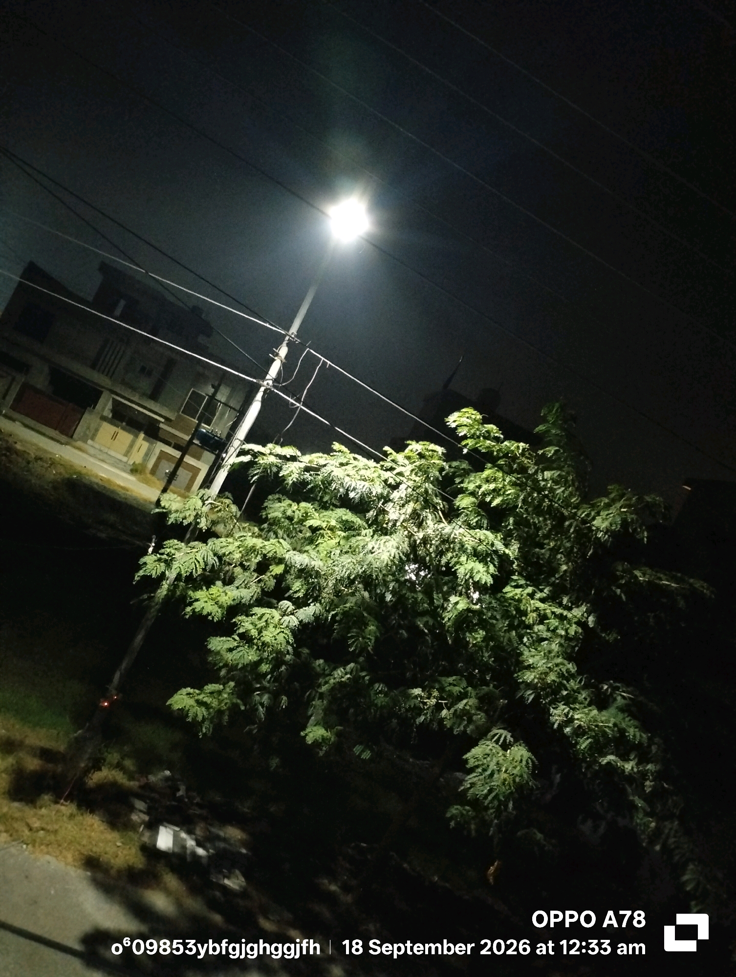 The image depicts a nighttime scene on a street in Badukke, Pakistan, illuminated by a streetlamp. A large, leafy tree occupies the right half of the frame, its foliage brightly lit by the streetlamp positioned on a utility pole on the left. Behind the tree and pole, a multi-story building is visible, its windows dark and its facade illuminated by the same light source. Power lines crisscross the sky above. At the bottom right of the image, the text "OPPO A78 18 September 2026 at 12:33 am" and a string of alphanumeric characters are overlaid, indicating the device and time the photo was taken. The ground in the foreground consists of grass and a paved area, with some debris scattered on the ground near the utility pole. No people are visible in the frame.