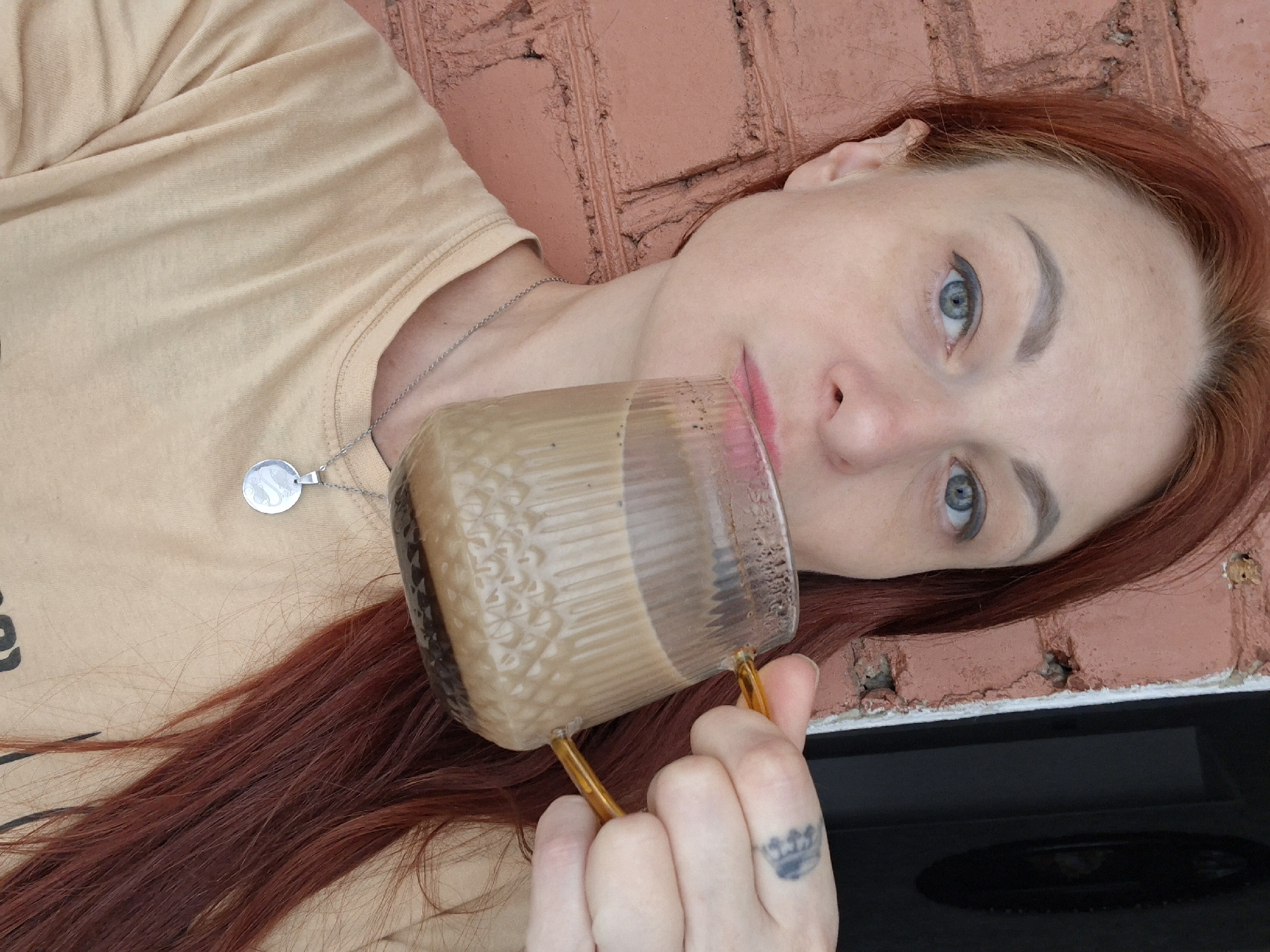 A close-up selfie captures a woman with auburn hair and blue eyes drinking from a glass mug filled with a light brown beverage. She is wearing a light tan t-shirt and a silver pendant necklace. A brick wall forms the background, with a dark doorway visible to the right. The woman's left hand is holding the mug, and a tattoo is visible on her index finger. The image appears to have been taken outdoors. The location is the city of Myadzyel, Belarus.