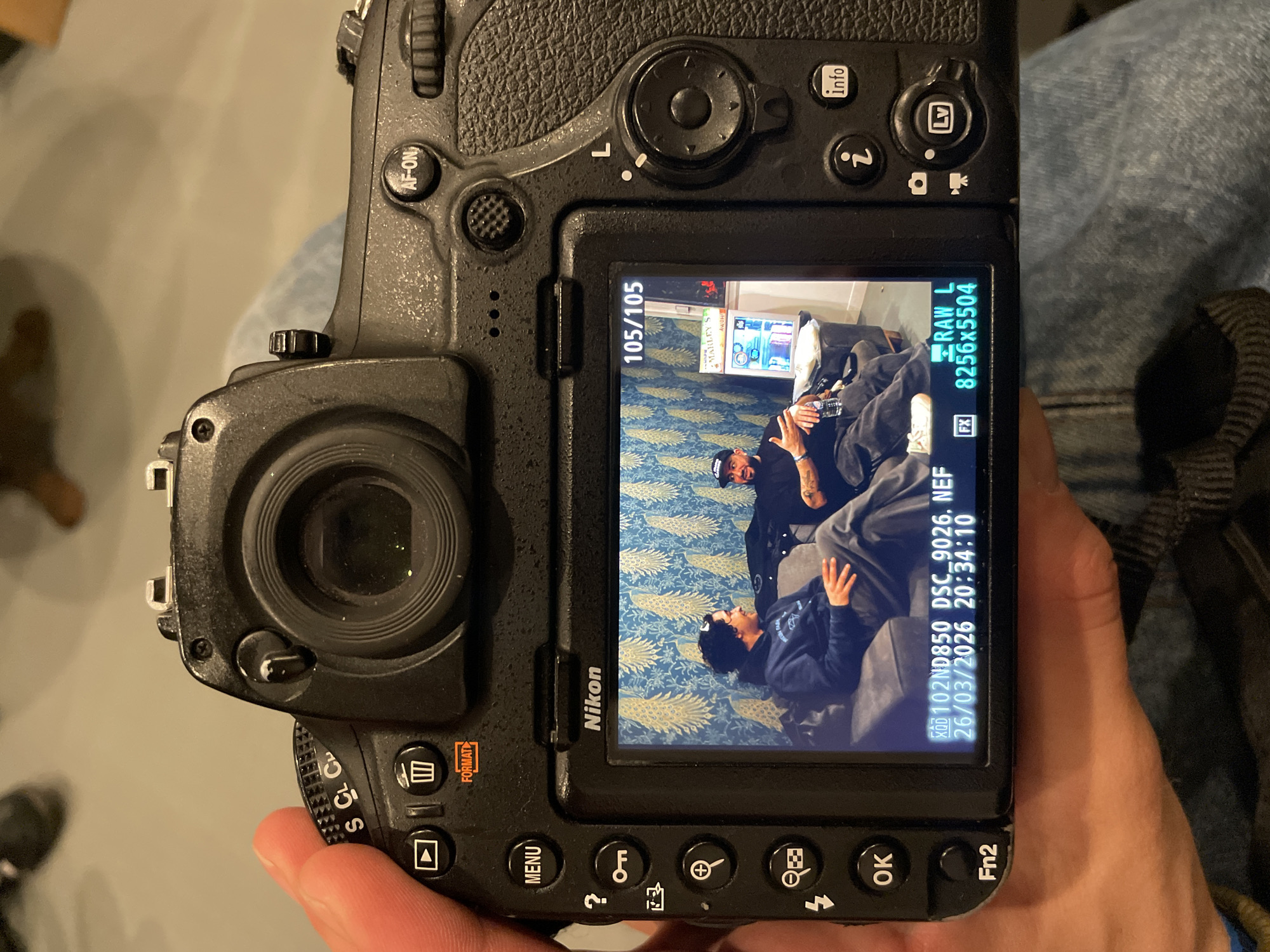 A person holds a black Nikon D850 DSLR camera, occupying the majority of the frame. The camera's rear LCD screen displays a captured image of two adult males seated on a dark grey sofa within an indoor setting.
The man on the left, wearing a dark hooded sweatshirt and grey pants, is looking rightward with a slight smile. The man on the right, clad in a black t-shirt, a black baseball cap, and black pants, is gesturing with his hands while holding a clear plastic water bottle. He exhibits tattoos on his forearms. Behind the men, a wall features blue wallpaper patterned with yellow and gold peacock feathers. To their right, a white cabinet or mini-fridge holds various items, including a box prominently labeled "MARLEY'S."
Overlayed text on the camera screen indicates the displayed image is "105/105" in a sequence, captured as a "RAW L" file with dimensions "8256x5504," identified by the filename "102ND850 DSC_9026.NEF." The capture date is "26/03/2026" at "20:34:10."
The depicted indoor scene, an unspecified location, is situated within the city of Paris, France. The individual holding the camera wears blue denim jeans, visible in the lower right portion of the image.